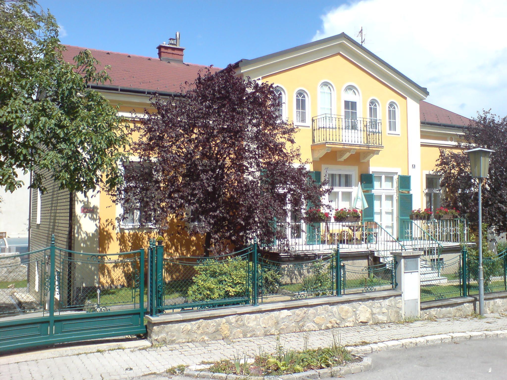 Žltý dom s balkónom a zeleným plotom, obklopený stromami.
