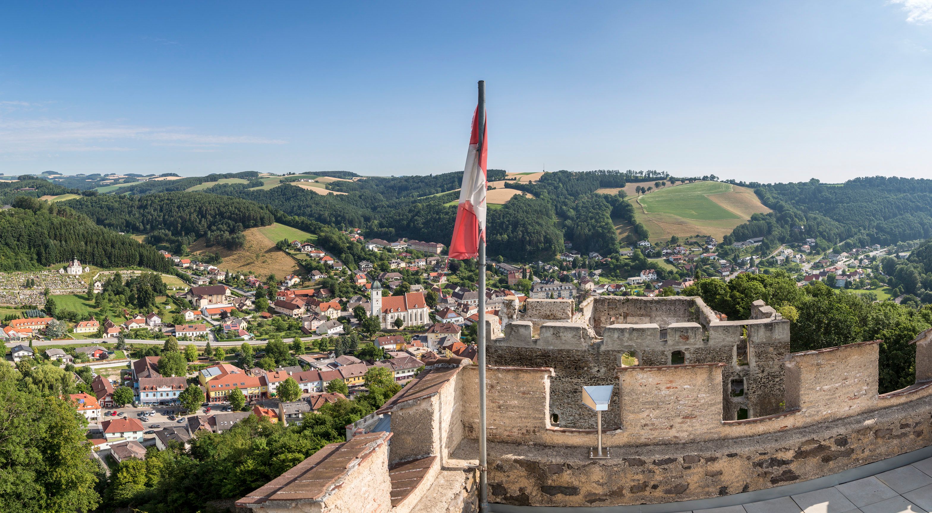 Panoramatický pohľad zo zrúcaniny hradu na mesto s kopcami v pozadí a rakúskou vlajkou v popredí.