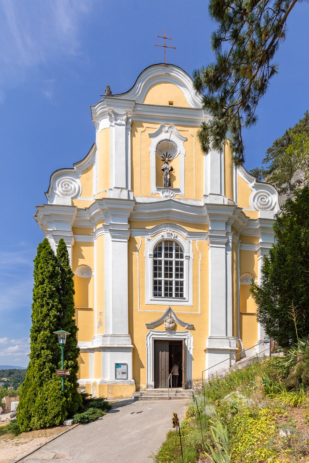 Žltý barokový kostol s bielymi stĺpmi a sochou nad vchodom, obklopený stromami a modrou oblohou.