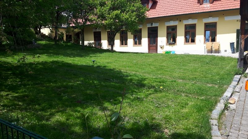 Zelená záhrada pred žltou budovou s červenými škridlami a niekoľkými oknami.