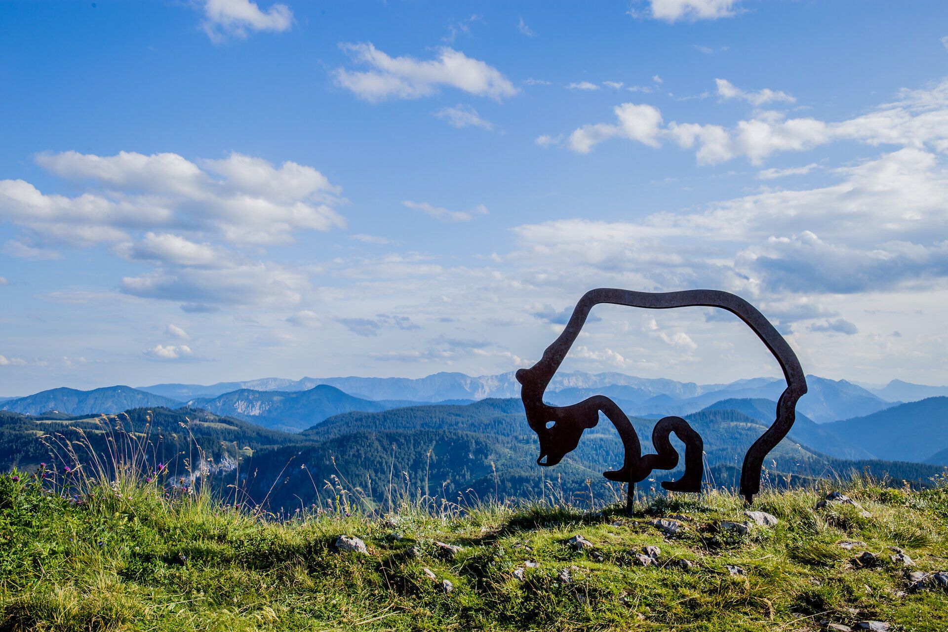 Ein majestätischer Blick auf die sanften Hügel und die beeindruckenden Berge, die sich unter einem strahlend blauen Himmel erstrecken. Die harmonische Verbindung von Natur und Kunst wird durch die stilvolle Skulptur des Ötscherbären unterstrichen, die den Wanderern ein Gefühl von Abenteuer und Freiheit vermittelt.