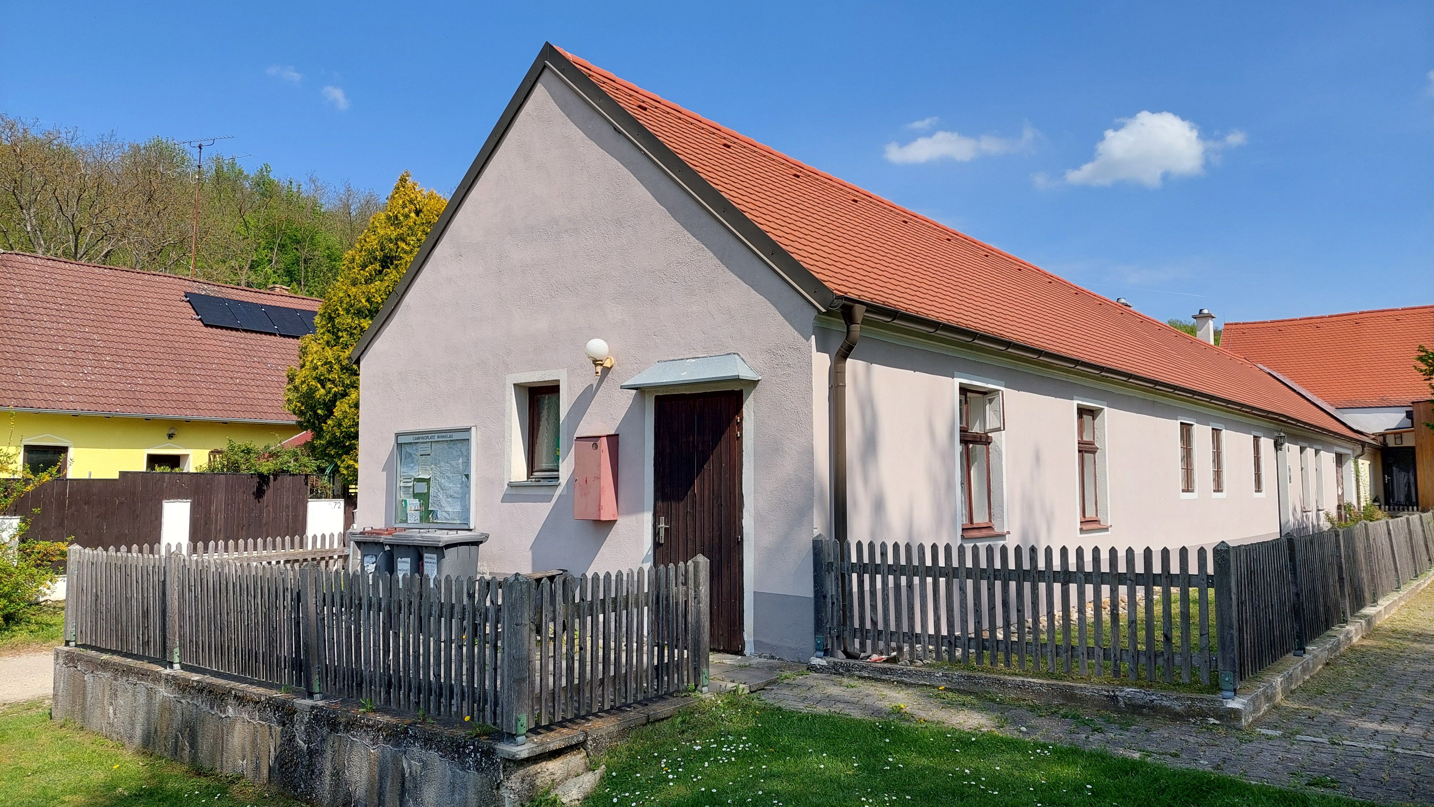 Dlhý jednoposchodový dom s červenou strechou a dreveným plotom, obklopený zelenou trávou a modrou oblohou.