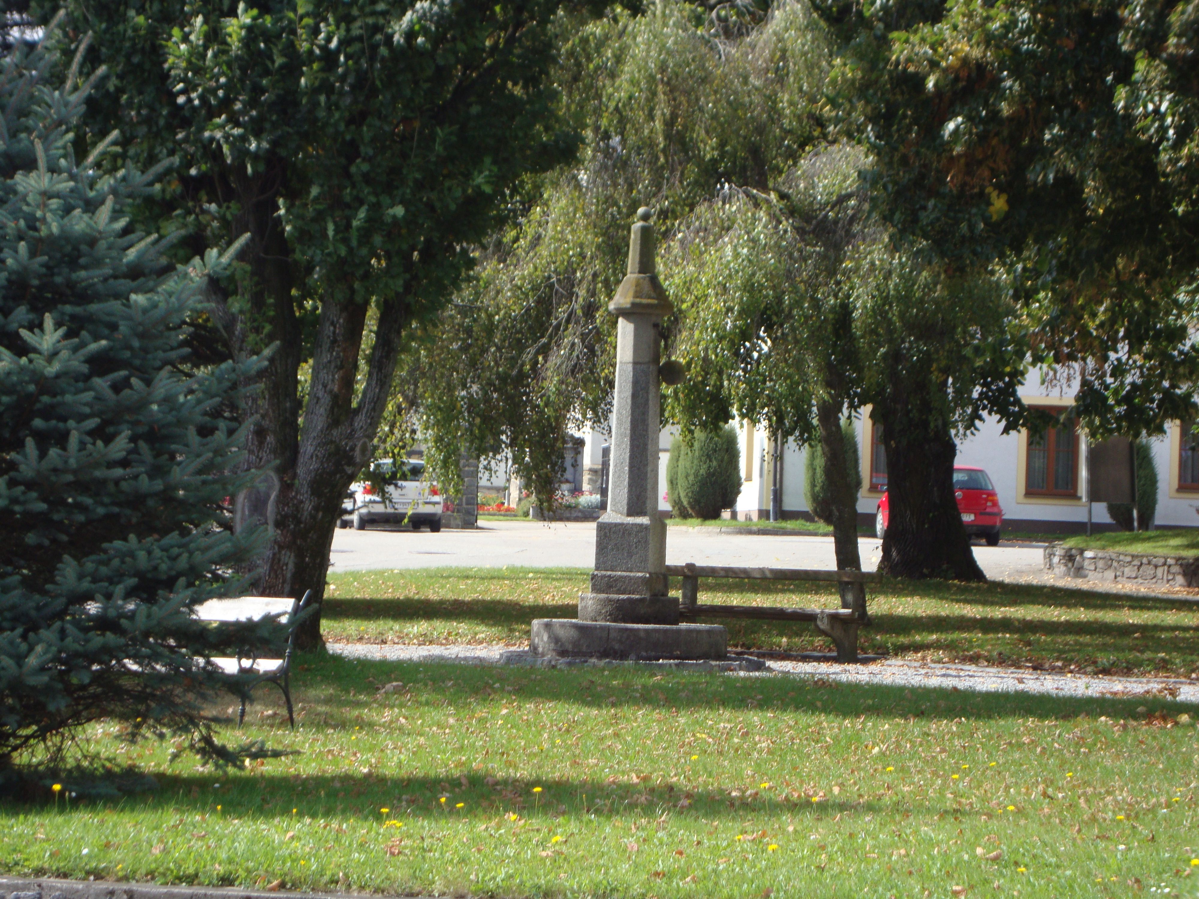 Malý park s kamenným pomníkom a lavičkou obklopený stromami.