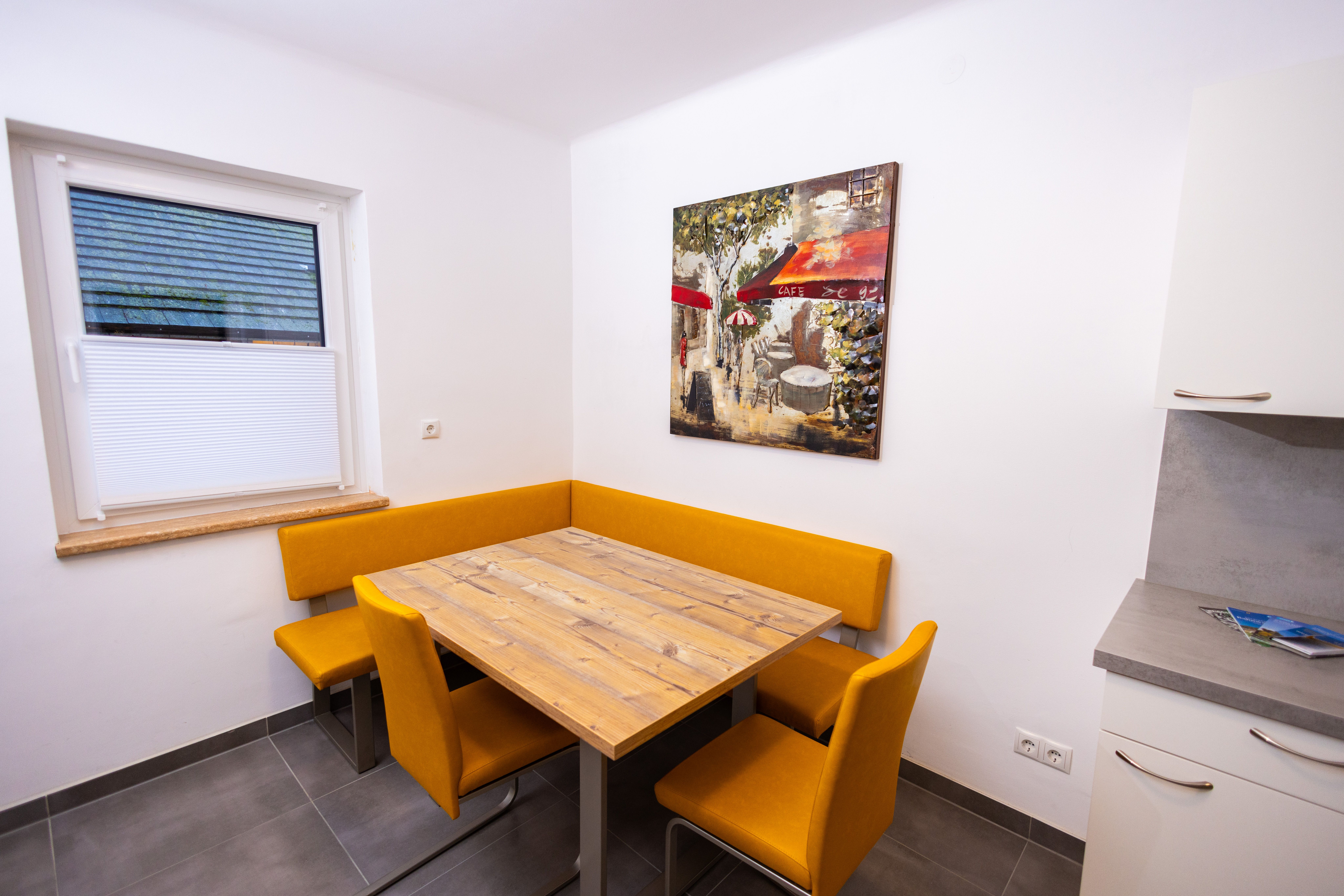 Jedálenský kút s dreveným stolom, žltými stoličkami a nástennou maľbou kaviarne.