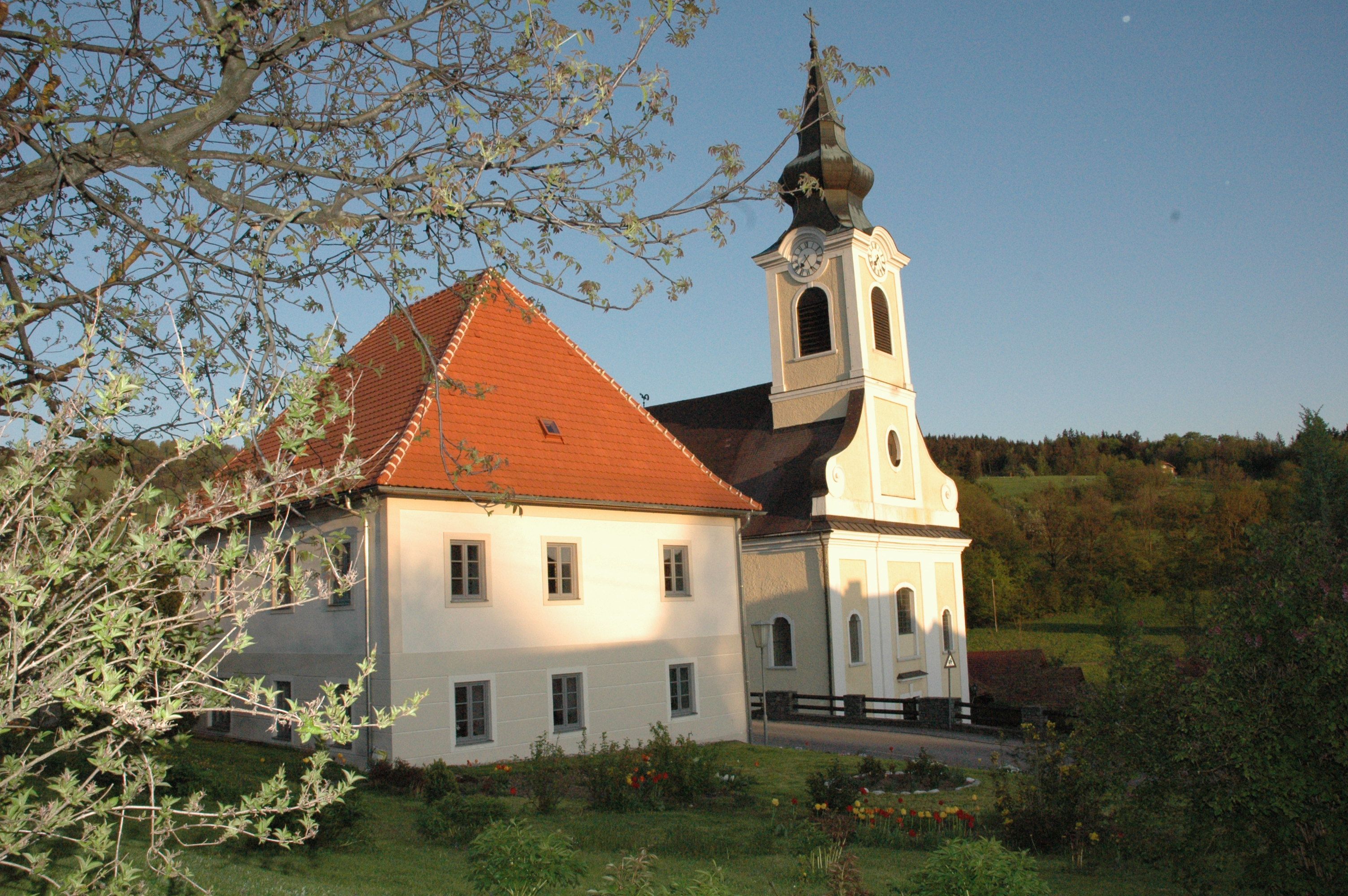 Kostol s farou vo vidieckom prostredí, obklopený stromami a lúkami.