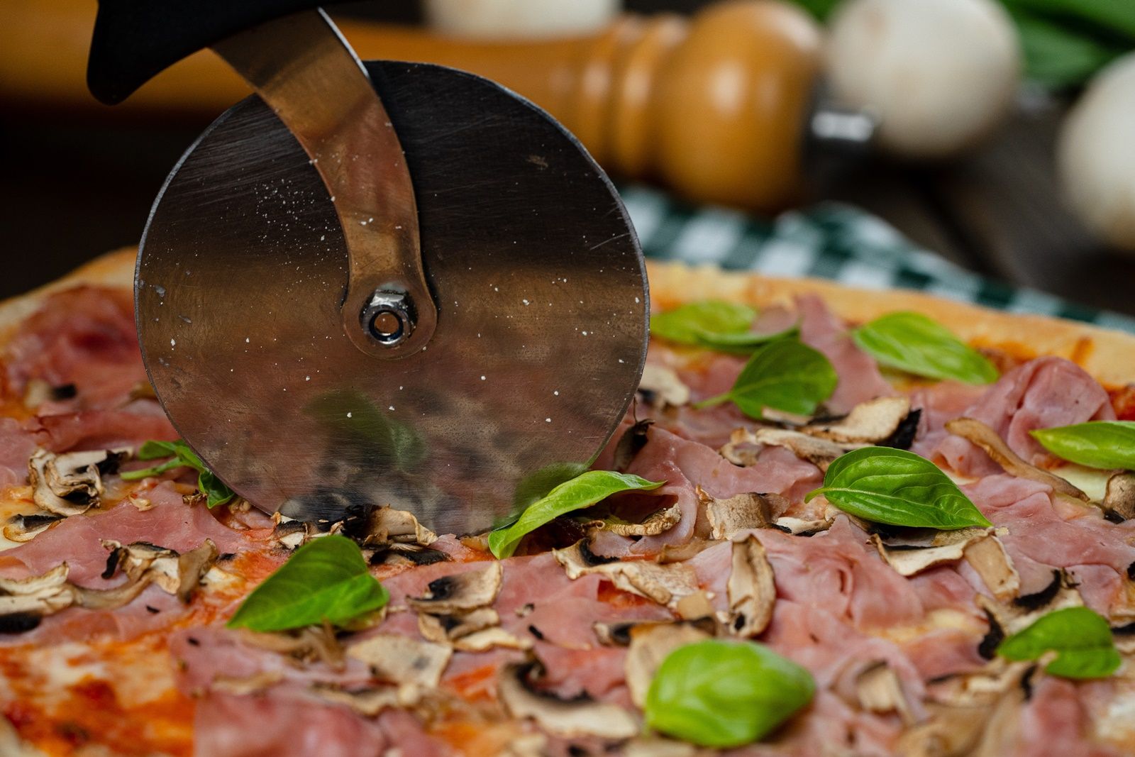 Detailný záber na pizzu so šunkou, šampiňónmi a bazalkou, ktorá sa krája nožom na pizzu.