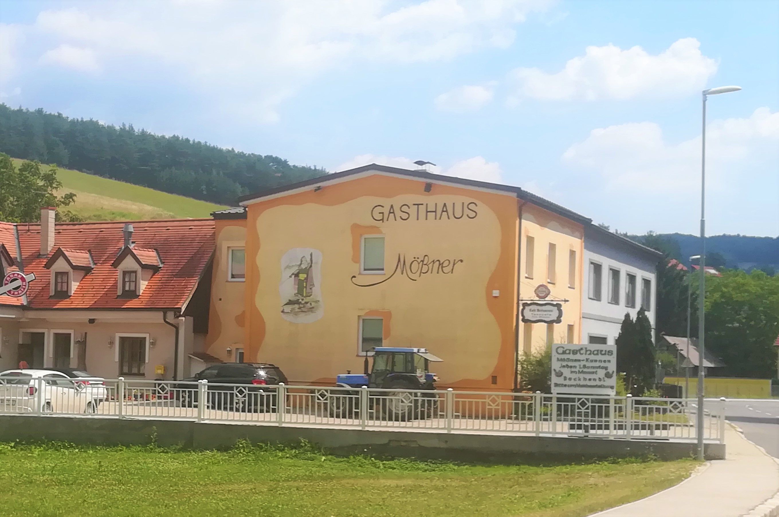 Exteriérový pohľad na žltooranžovo natretý hostinec Mößner vo vidieckom prostredí s modrým traktorom a autami pred budovou.
