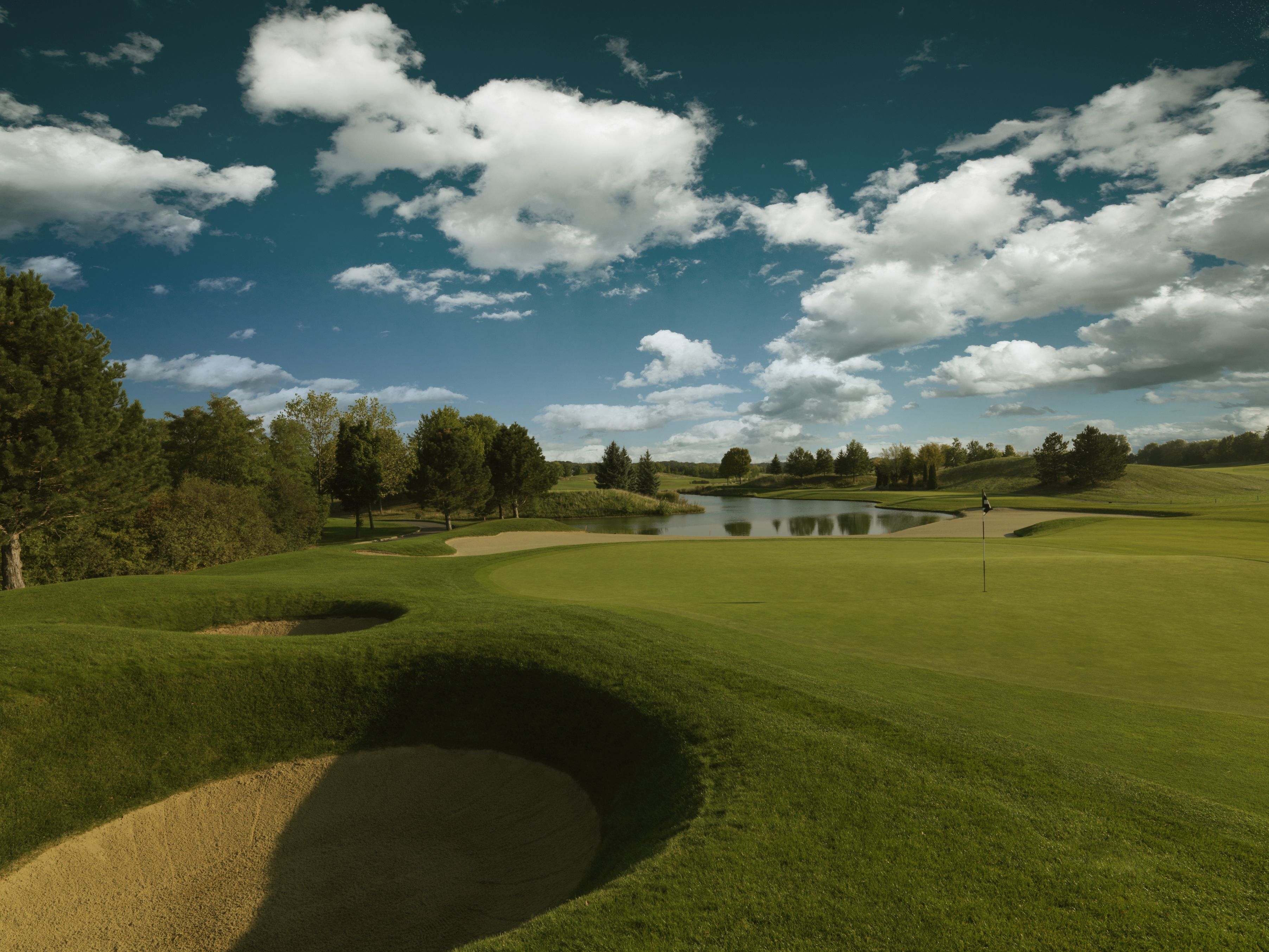 Golfové ihrisko s pieskovým bunkrom, zeleným a rybníkom pod modrou oblohou s mrakmi.