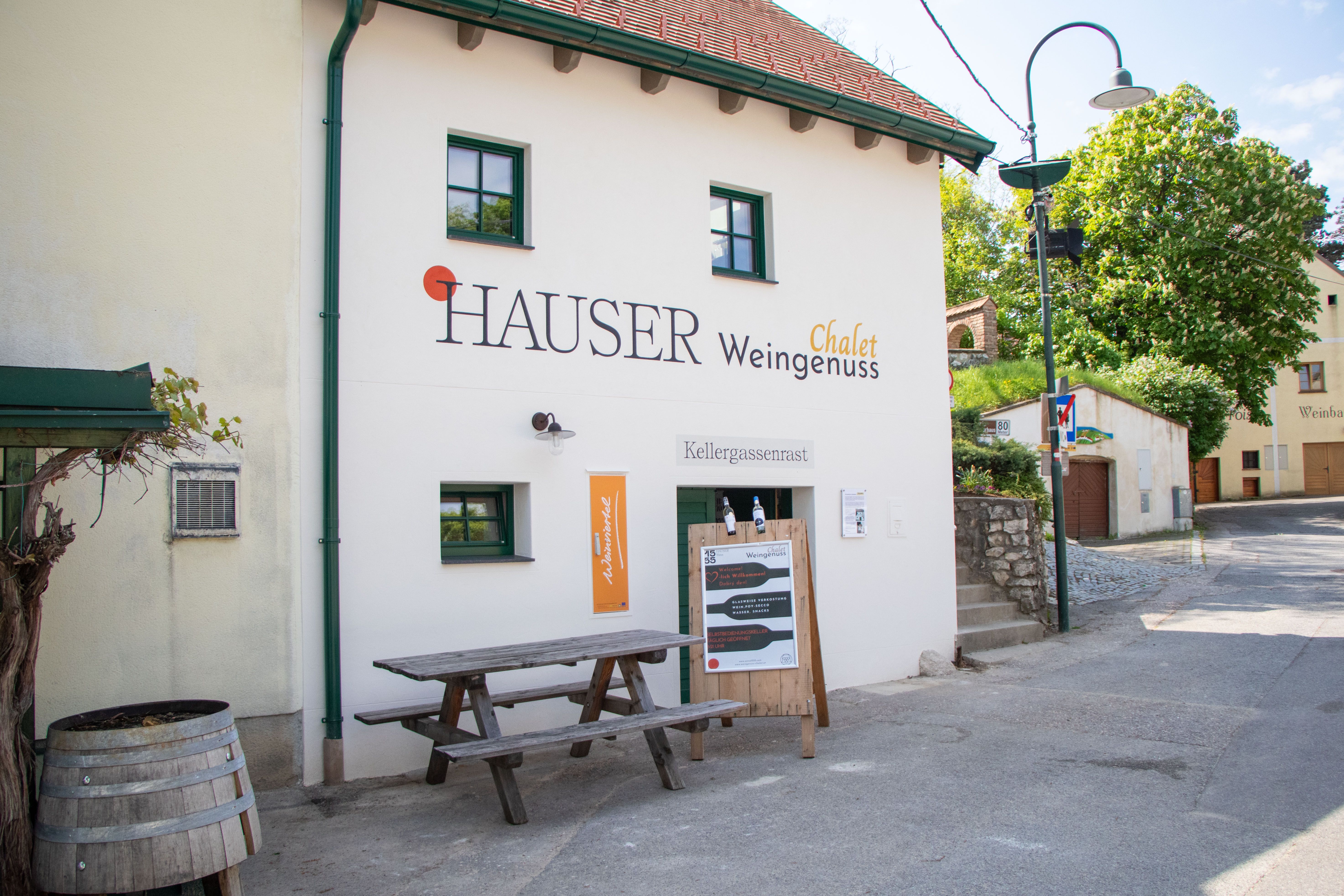 Pohľad na budovu s označením "Hauser Chalet Weingenuss" zvonku. Pred budovou je drevený stôl a sud na víno.
