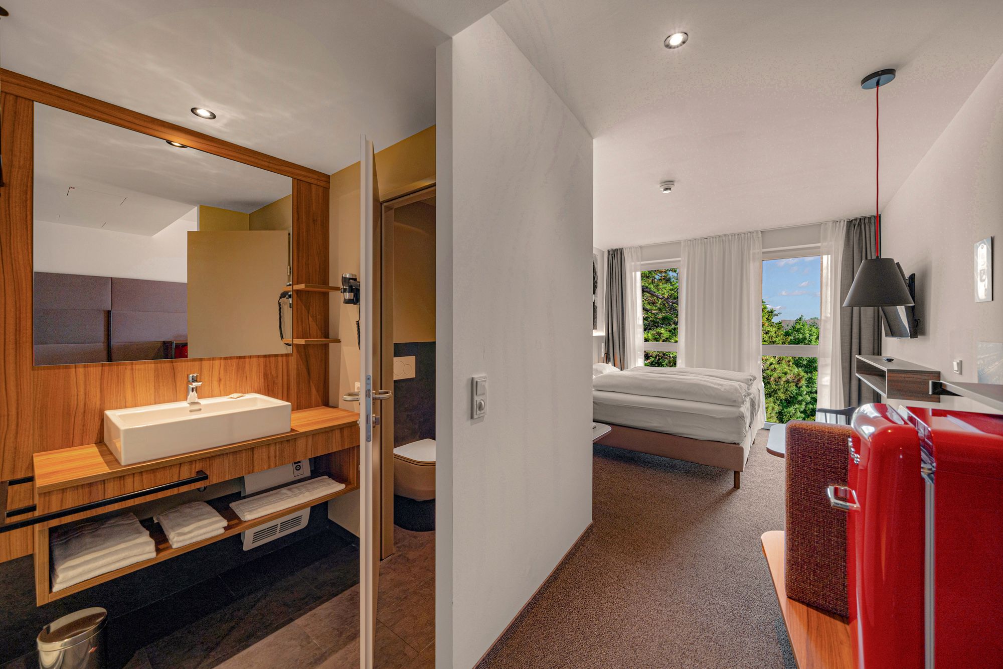 Pohľad do interiéru modernej hotelovej izby s kúpeľňou, posteľou a červenou chladničkou.