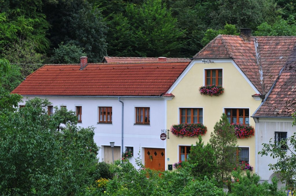 Tradičný dom s červenými škridlami a kvetinovými truhlíky pred oknami, obklopený zelenou vegetáciou.