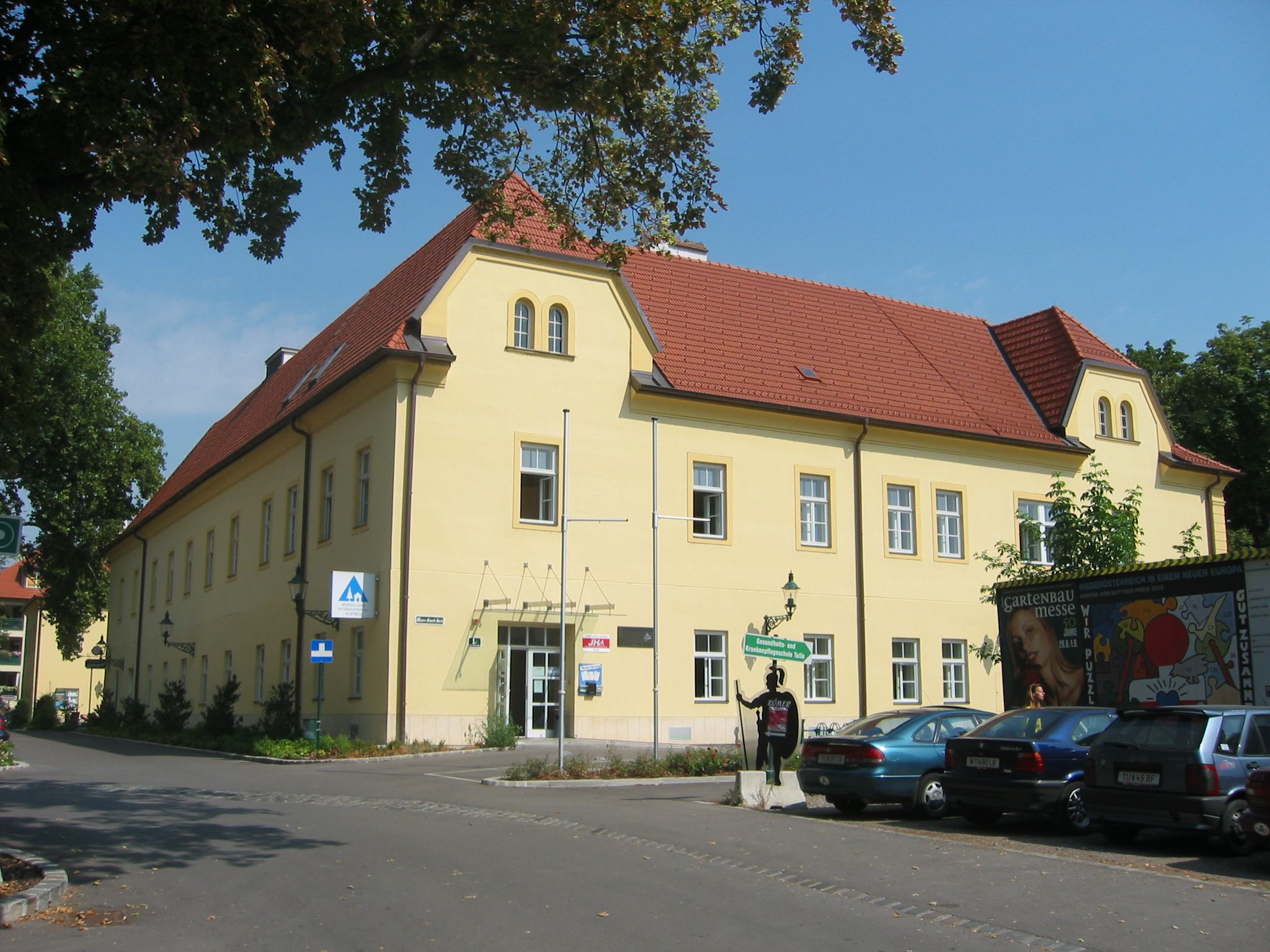Exteriérový pohľad na žltú budovu s červenou strechou, Junges Hotel Tulln, obklopenú stromami a zaparkovanými autami.