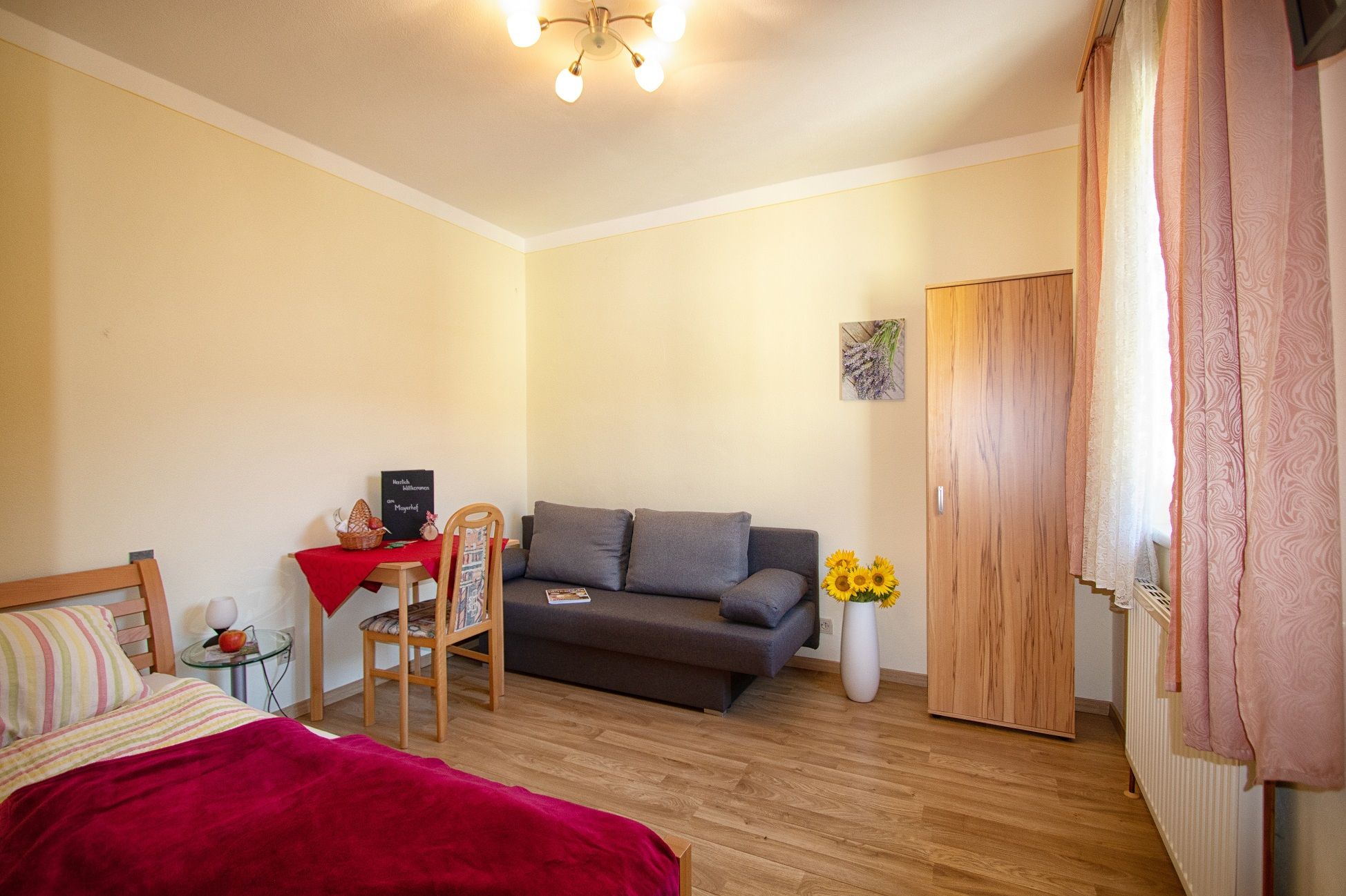 Útulná izba s posteľou, pohovkou, stolom a skriňou, zdobená slnečnicami a obrazom na stene.