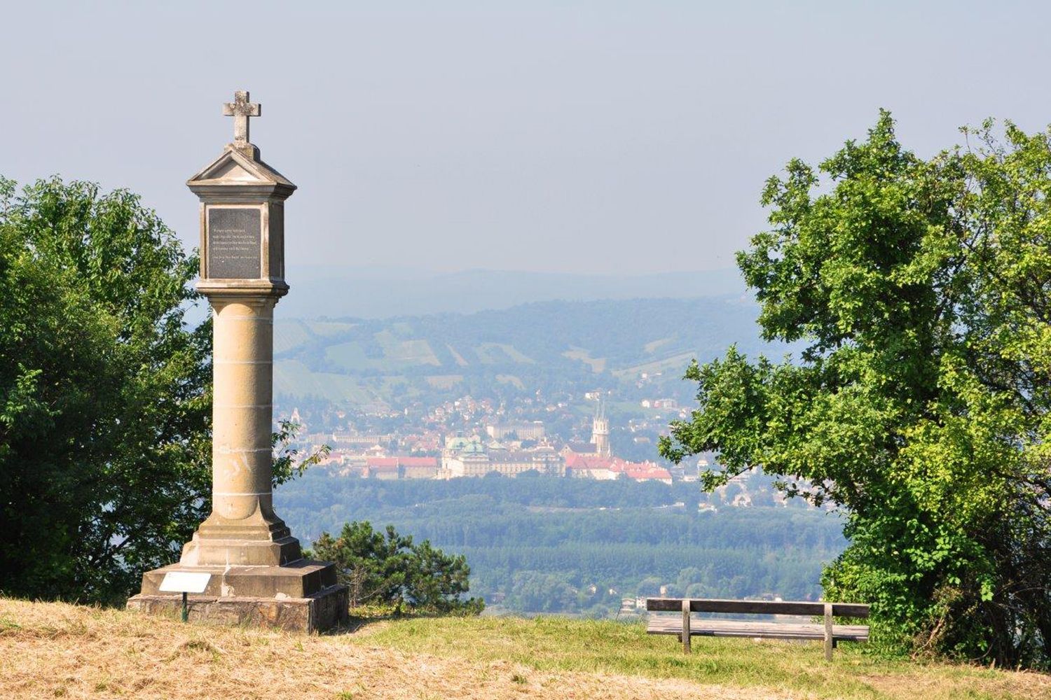 Stĺp s krížom na kopci, v pozadí mesto a kopce.