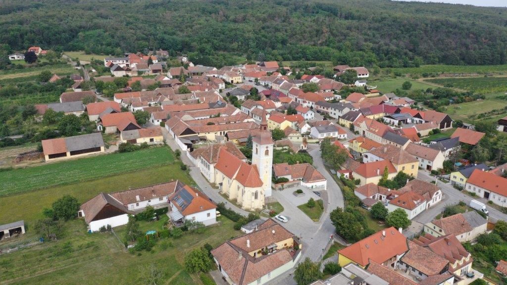 Letecký pohľad na malú dedinu s centrálnym kostolom a červenými strechami, obklopenú zelenými poliami a lesmi.