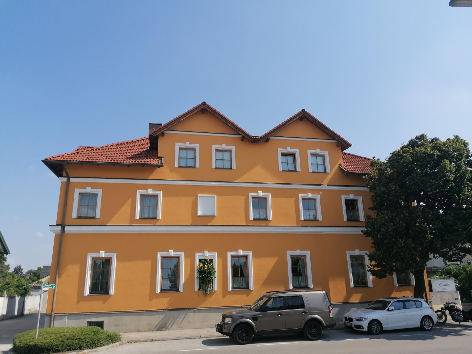 Žltá budova s červenými škridlami a niekoľkými oknami, pred ňou zaparkované autá a strom.