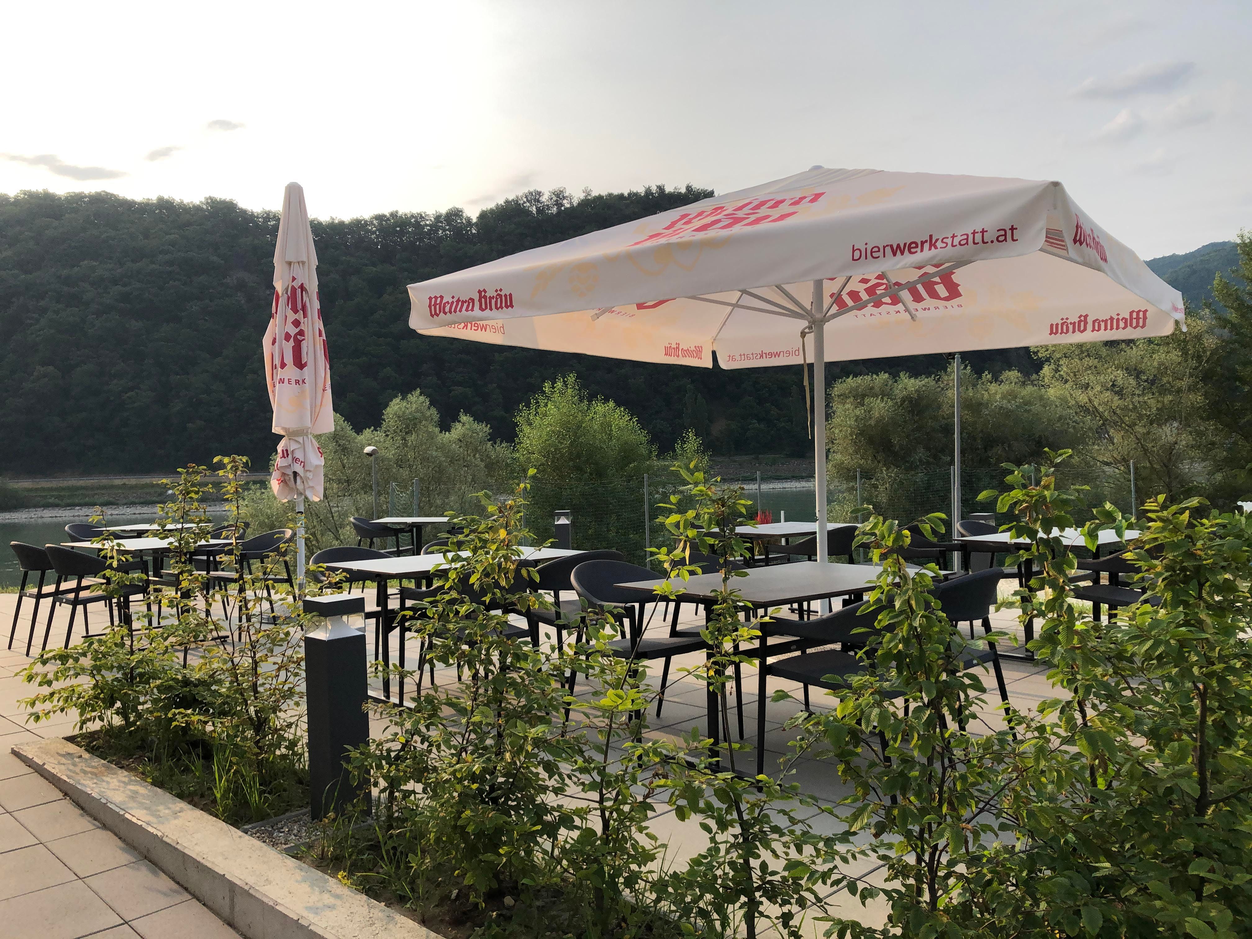 Terasa so stolmi, stoličkami a slnečníkmi, obklopená prírodou.