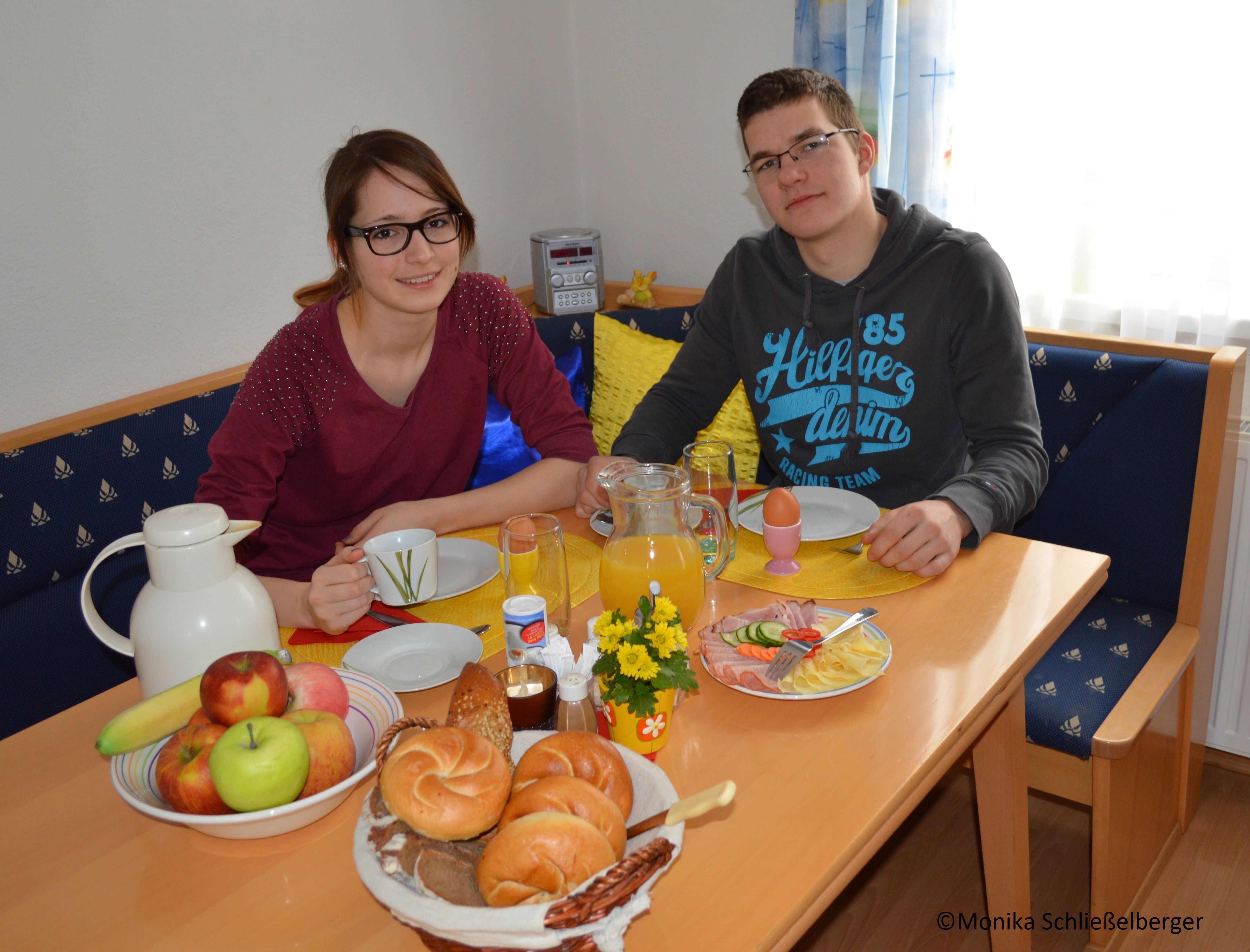 Dve osoby sedia pri stole s raňajkami, ktoré obsahujú pečivo, ovocie a džús.