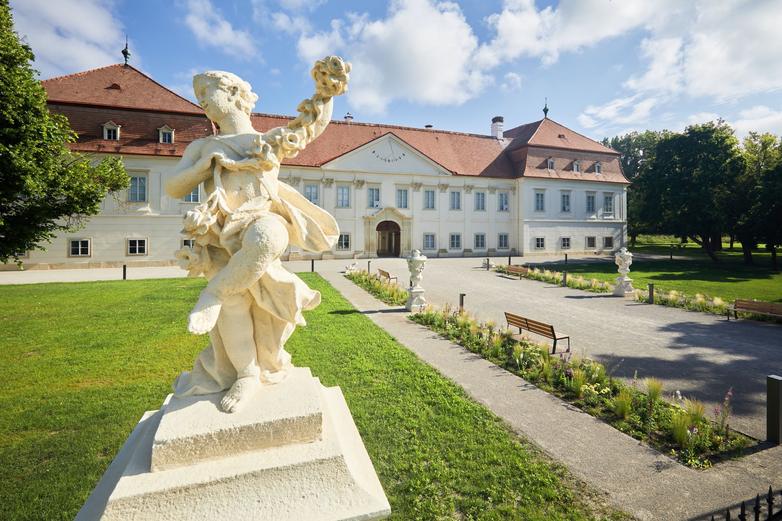 Baroková socha pred historickou budovou s červenou strechou a udržiavanou záhradou.