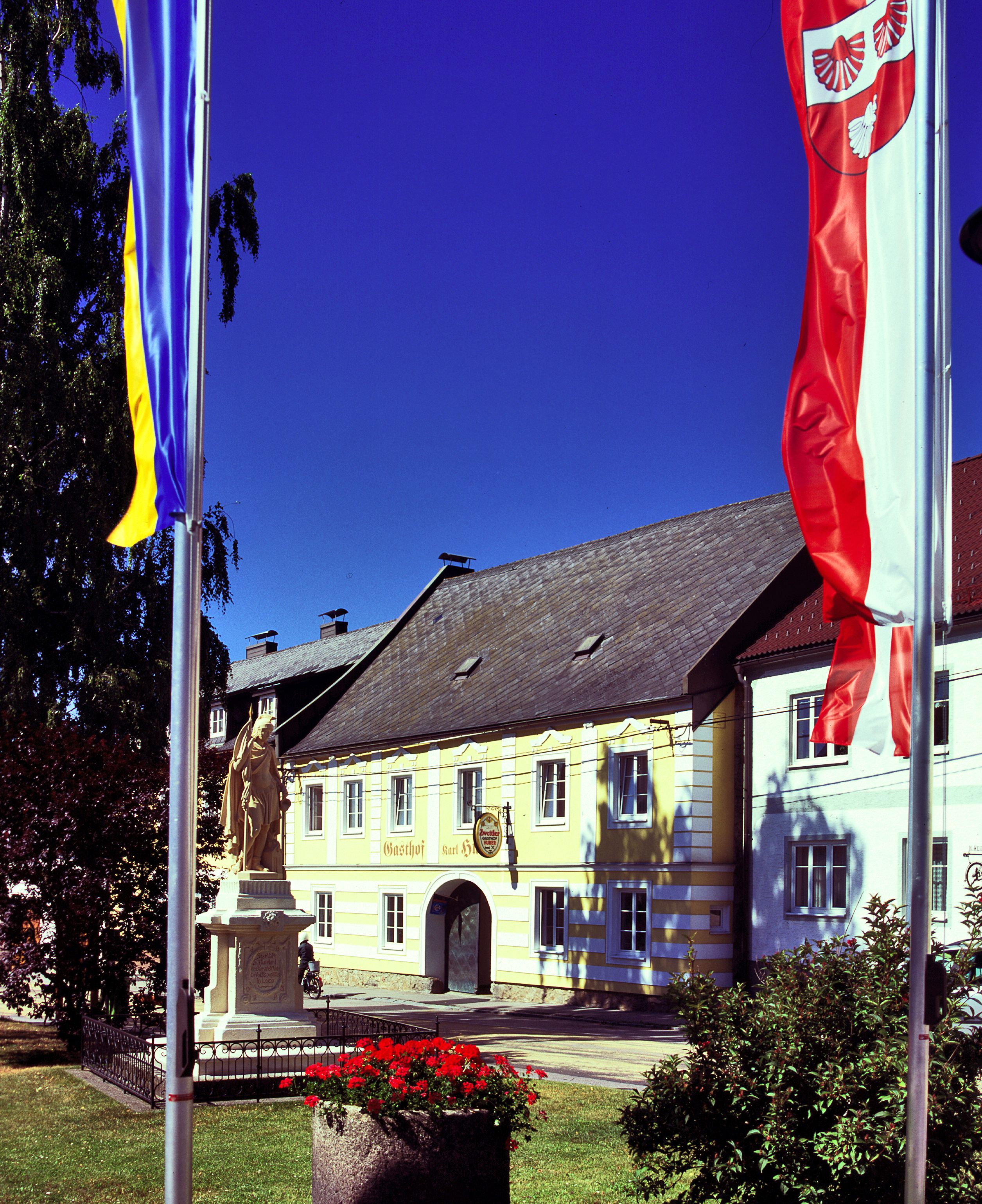 Žltá budova s nápisom "Gasthof Karl Huber", obklopená vlajkami a sochou.