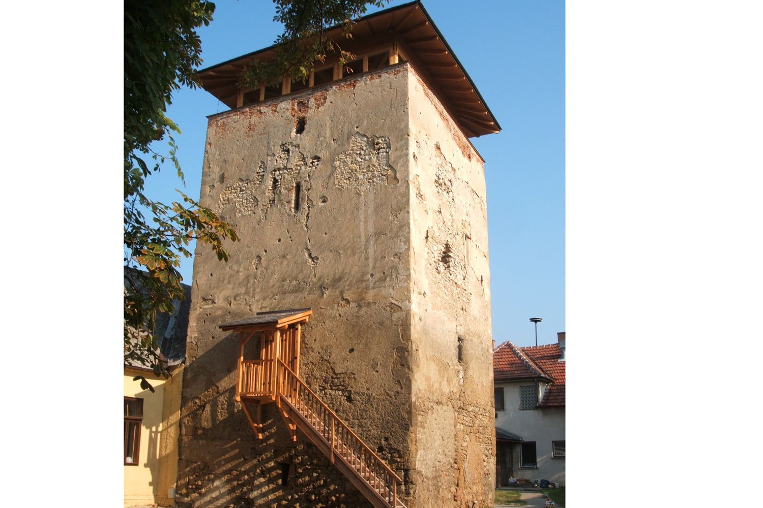 Stará obranná veža s rozpadajúcou sa fasádou a dreveným schodiskom, obklopená domami a stromami.