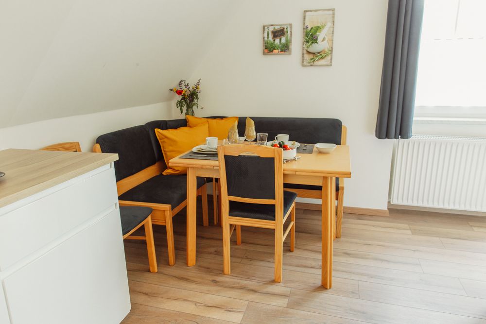 Jedálenský kút s dreveným stolom, stoličkami a rohovou lavicou, s riadom, kyticou kvetov a dekoráciou na stene.