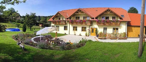 Veľký žltý dom s červenými strechami a kvetinovými truhlíky, obklopený udržiavanou záhradou s posedením a slnečníkom.