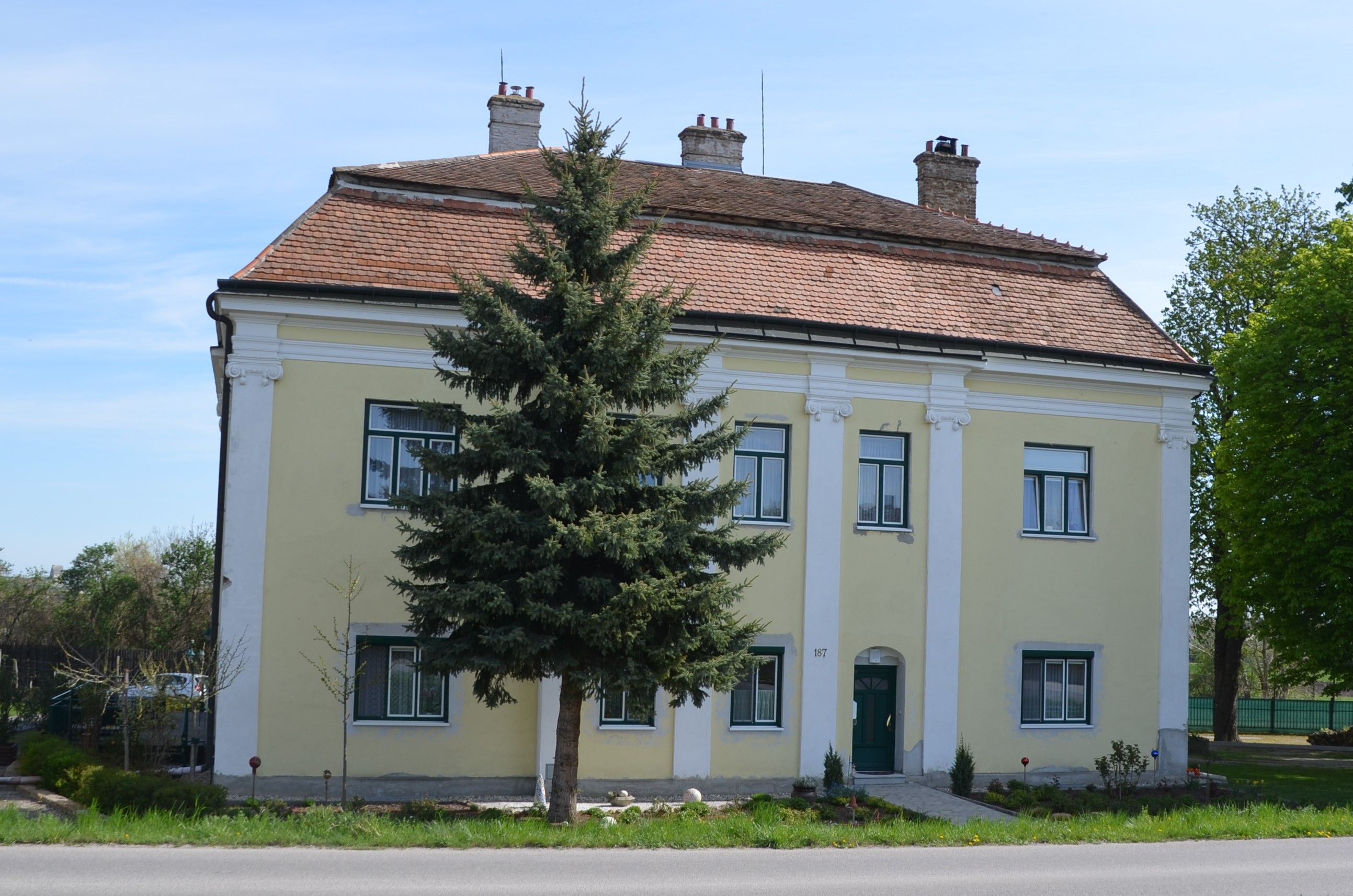 Žltá dvojpodlažná budova s červenou škridlovou strechou a zelenými dverami, obklopená stromami a malou záhradou.