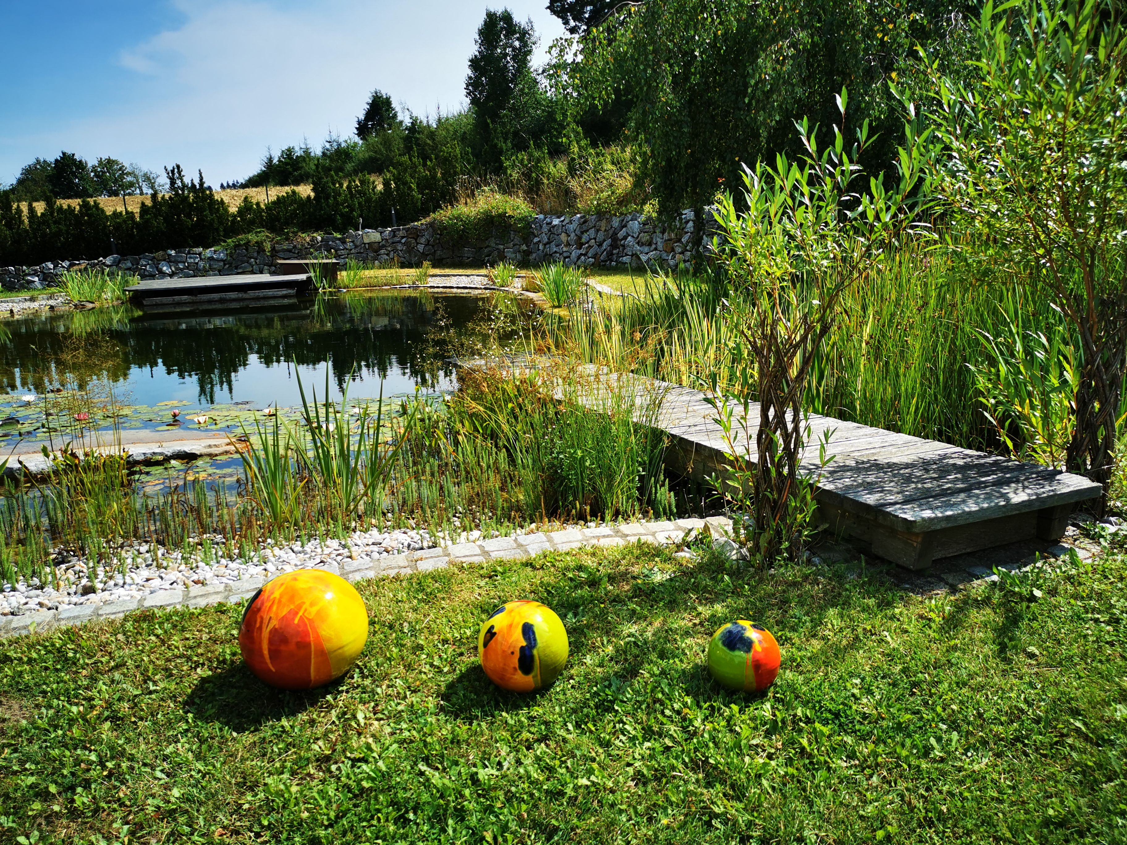 Prírodné kúpacie jazierko s dreveným chodníkom a farebnými loptami na trávniku.