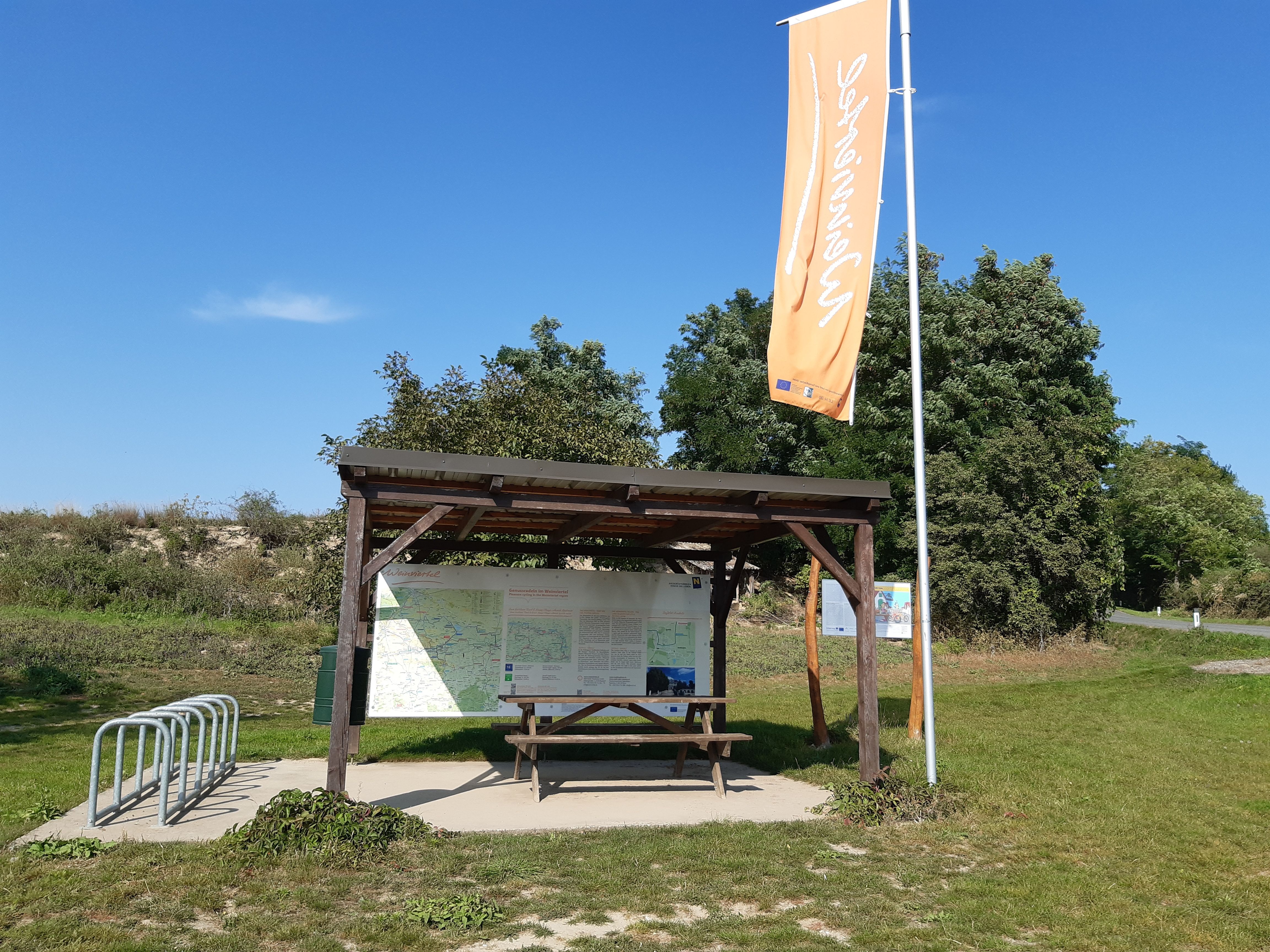 Odpočinková stanica s krytým stolom, stojanom na bicykle a informačnými tabuľami v zelenom prostredí.