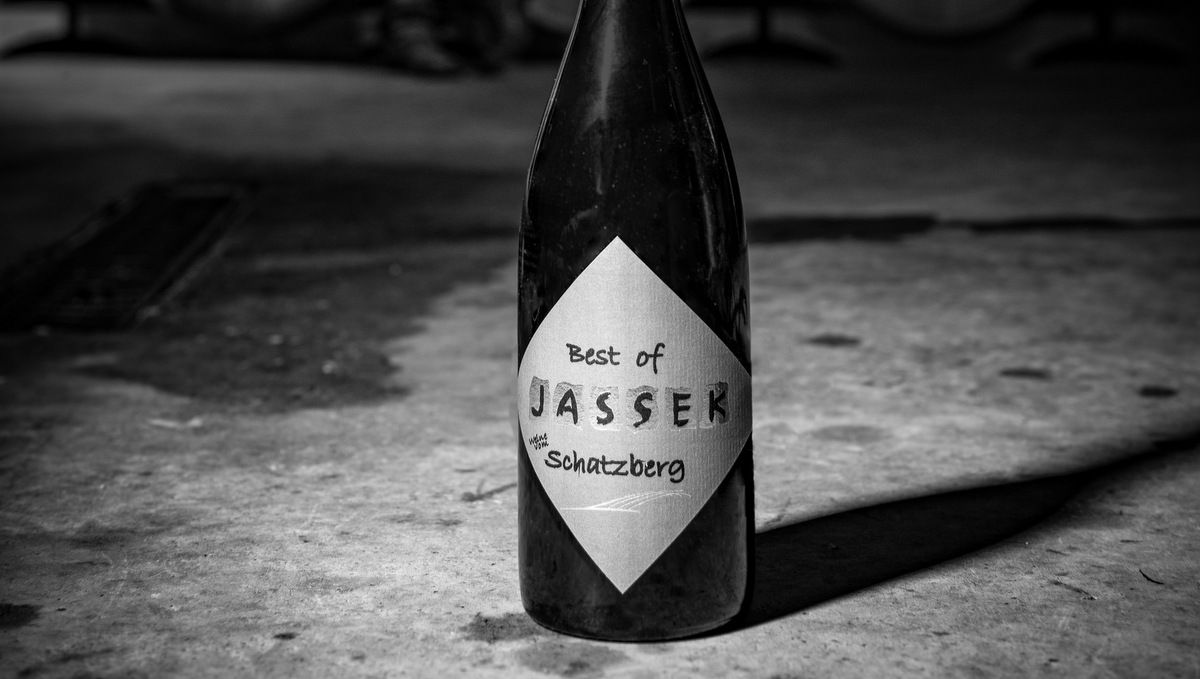 Čiernobiela fotografia fľaše vína s nápisom "Best of Jassek Schatzberg" na betónovej podlahe.