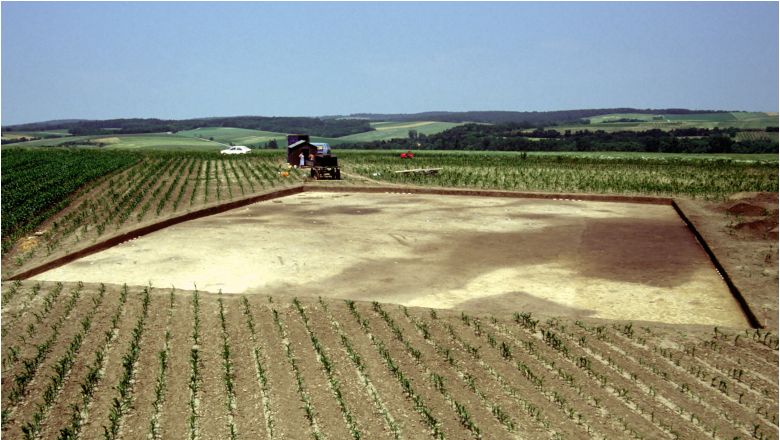 Archeologické vykopávky na poli s poľnohospodárskymi strojmi v pozadí.