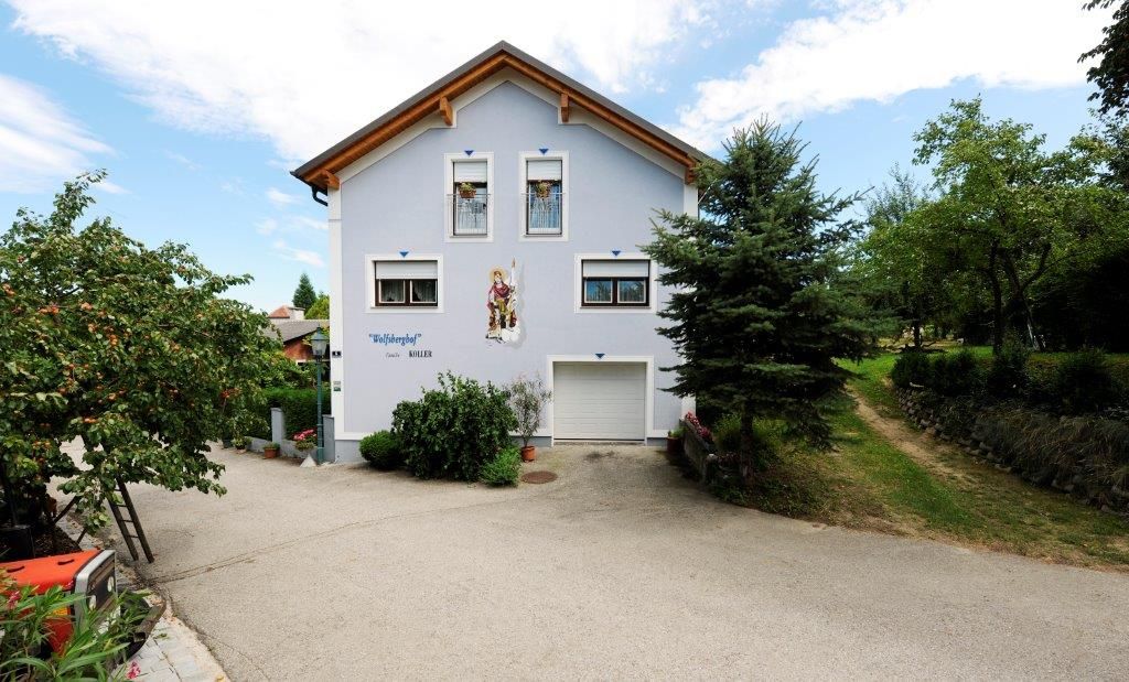 Dvojpodlažný dom s modrou fasádou a drevenou strechou, obklopený stromami a vydláždenou cestou.