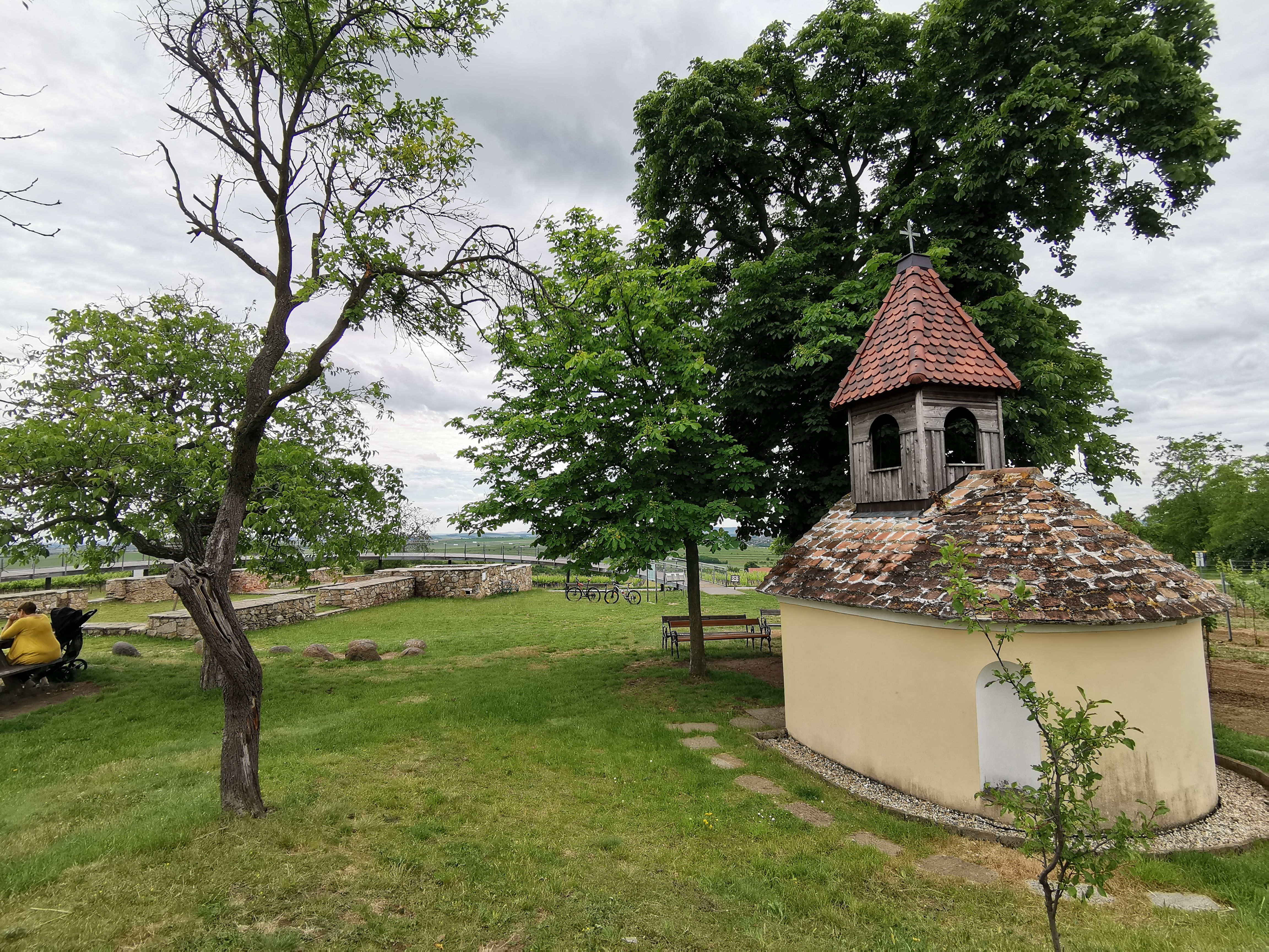 Malá kaplnka s červenou škridlovou strechou v zelenej krajine, obklopená stromami a lavičkami.