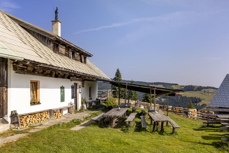Tradičná horská chata s drevenými stolmi a lavicami vonku, obklopená zelenými lúkami a zalesnenými kopcami pod modrou oblohou.