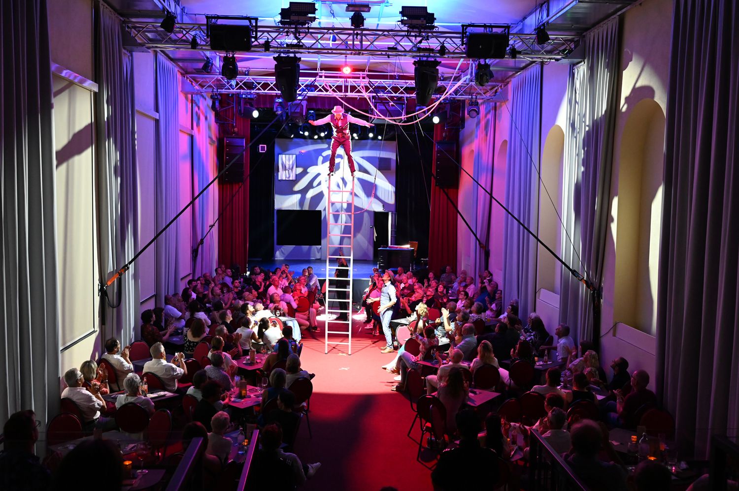 Umelec balansuje na rebríku v preplnenom divadle s farebným osvetlením.