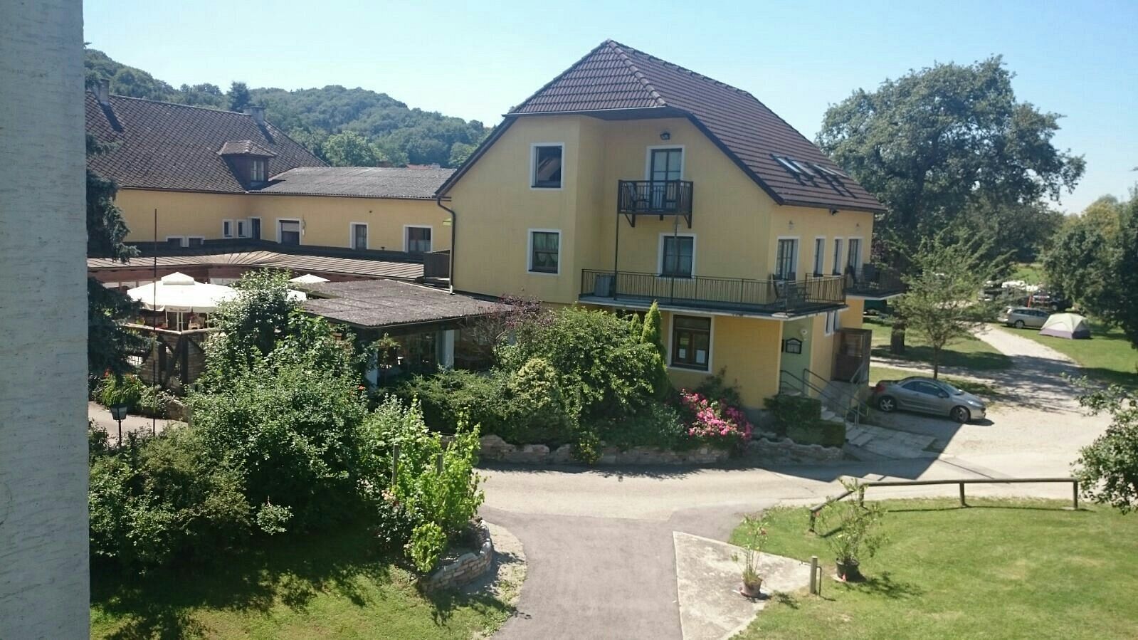 Žltá budova s terasou a záhradou, obklopená stromami a autom na parkovisku.