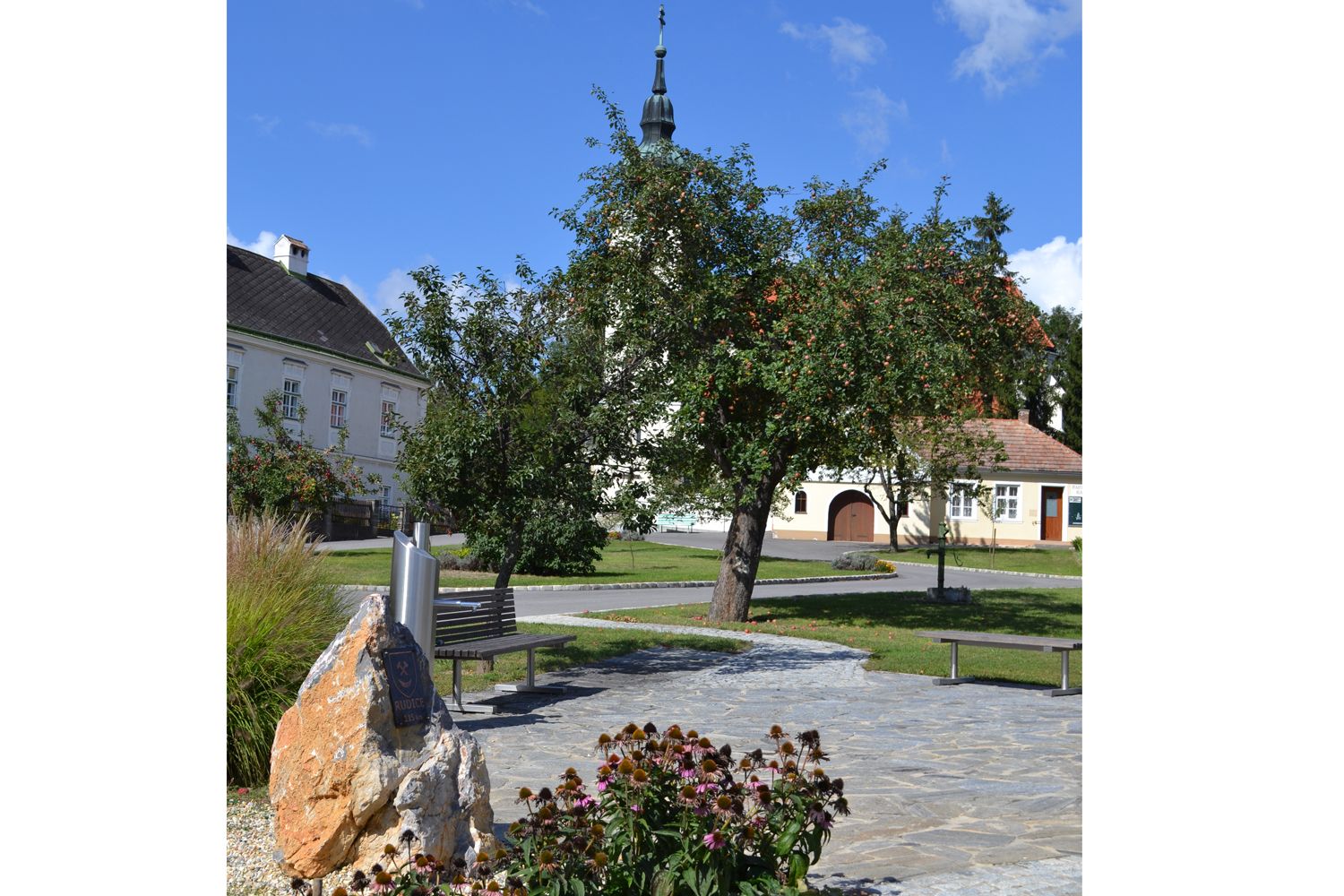 Dedinské námestie v Hagenbergu s kostolom, stromami a lavičkami.