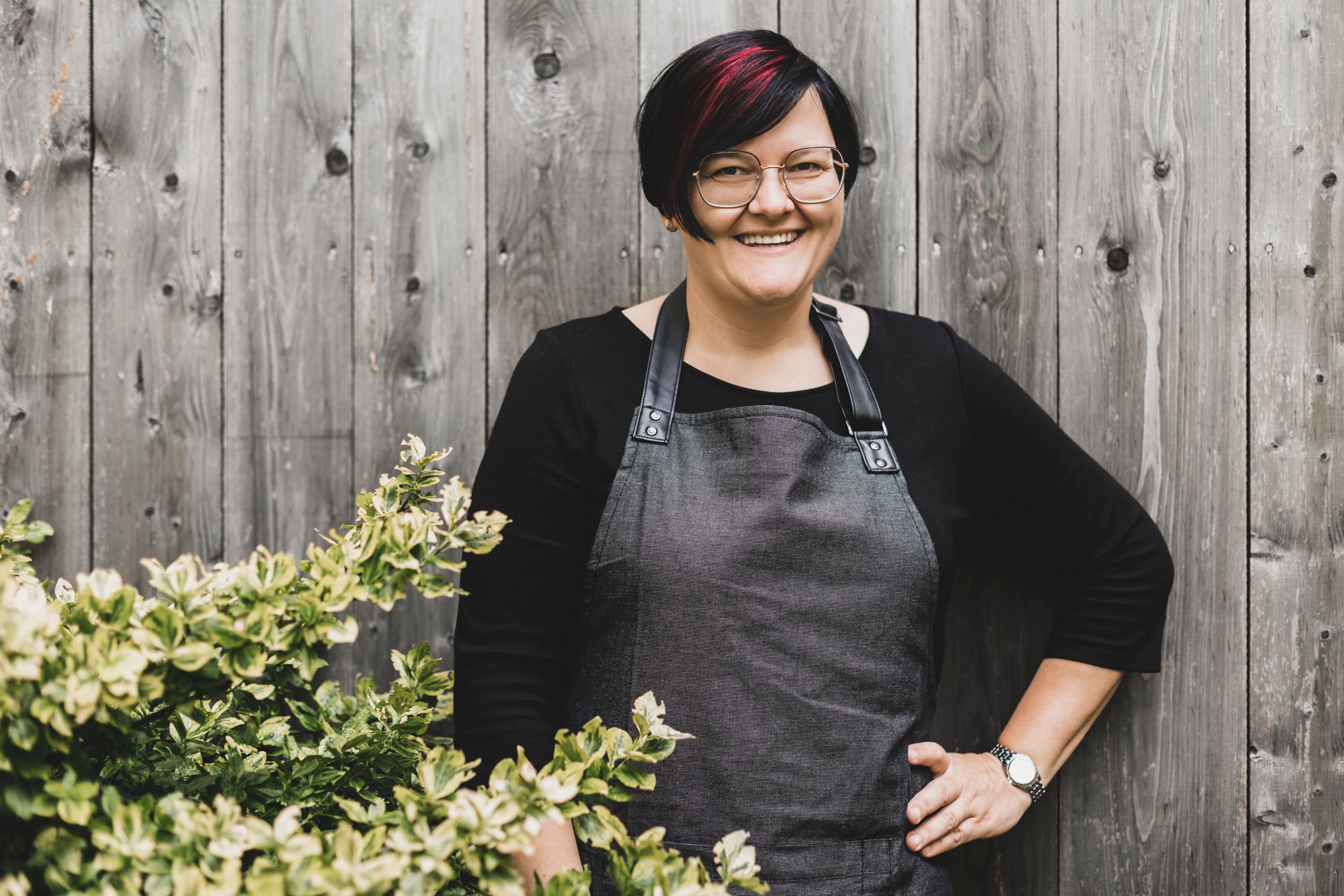 Žena v okuliaroch a zástere stojí s úsmevom pred drevenou stenou vedľa kríka.