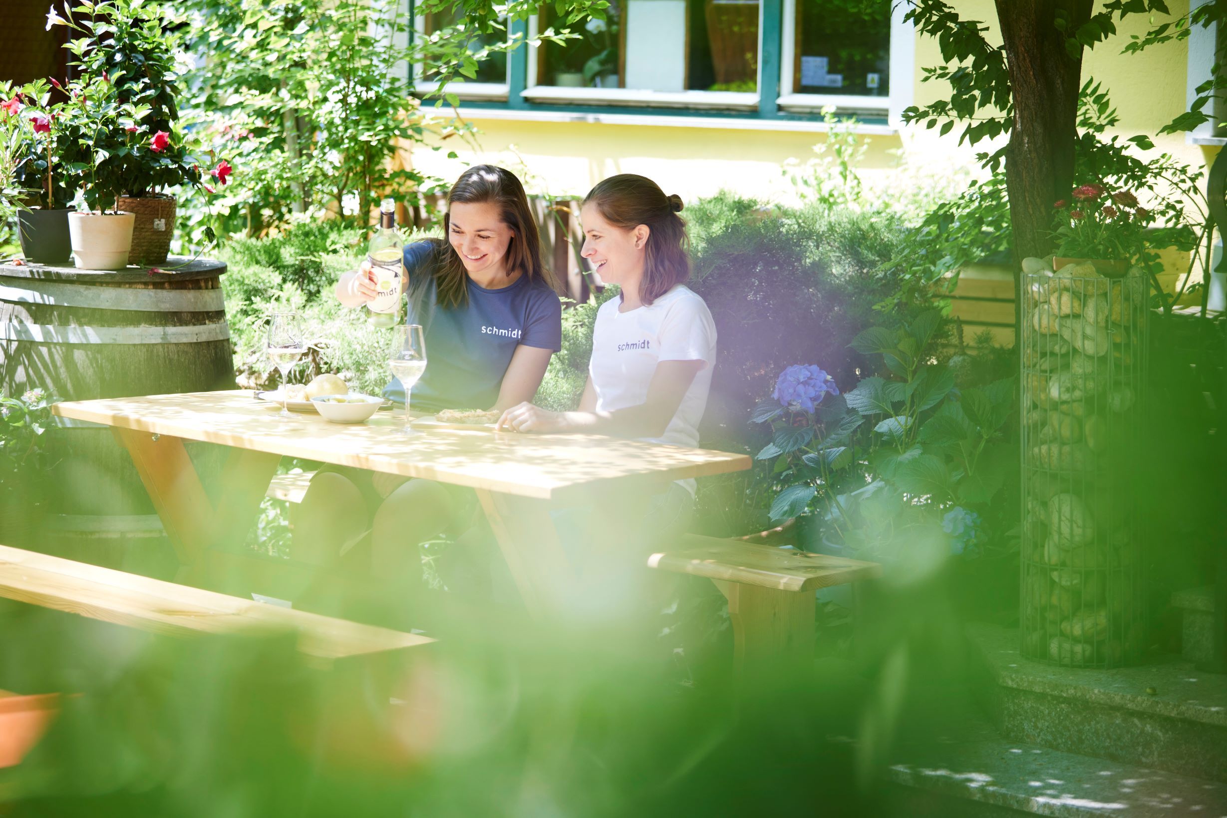 Dve ženy sedia pri drevenom stole v záhrade a vychutnávajú si víno.