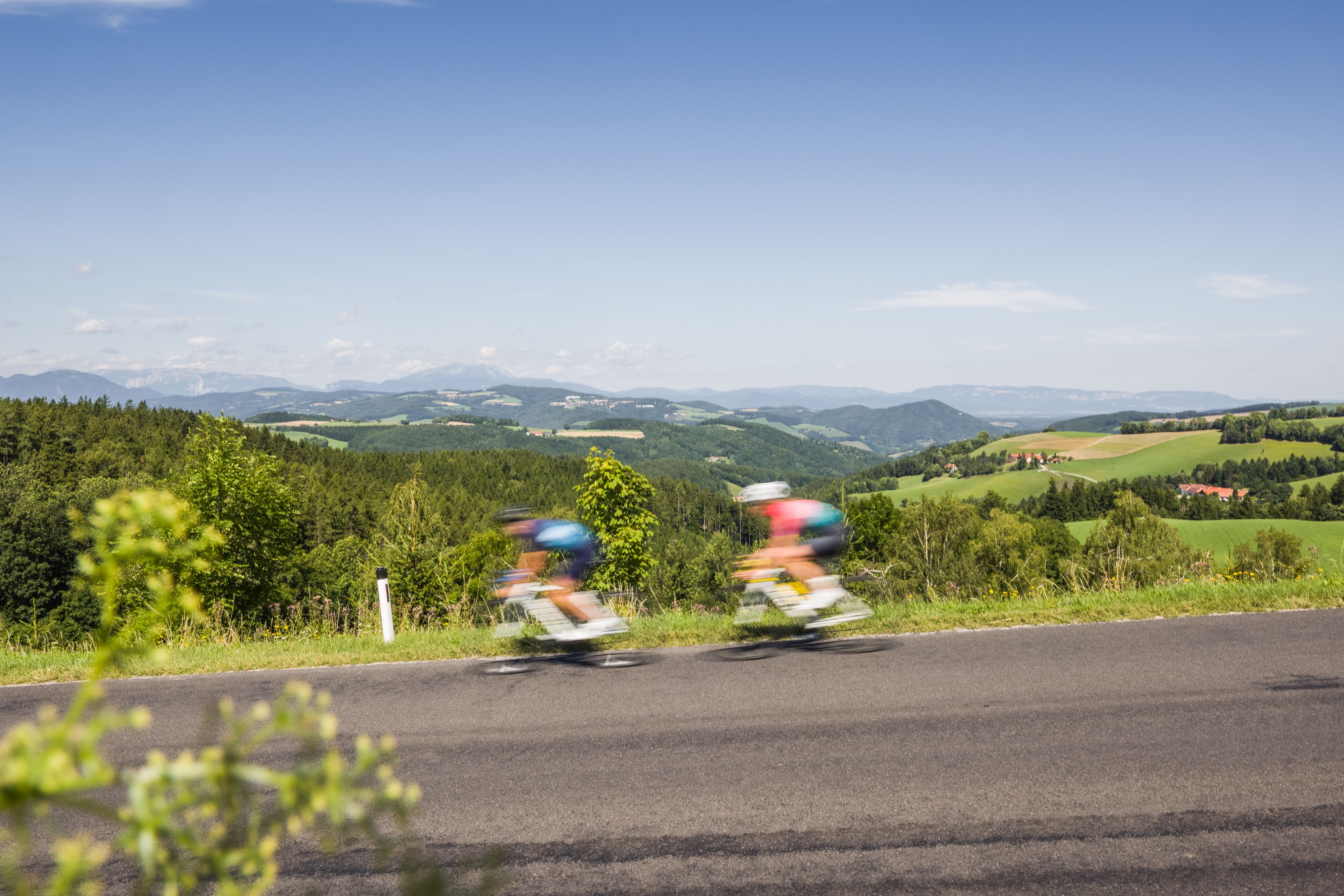 Dvaja ľudia na cestnom bicykli idú rýchlo po ceste v kopcovitej krajine.