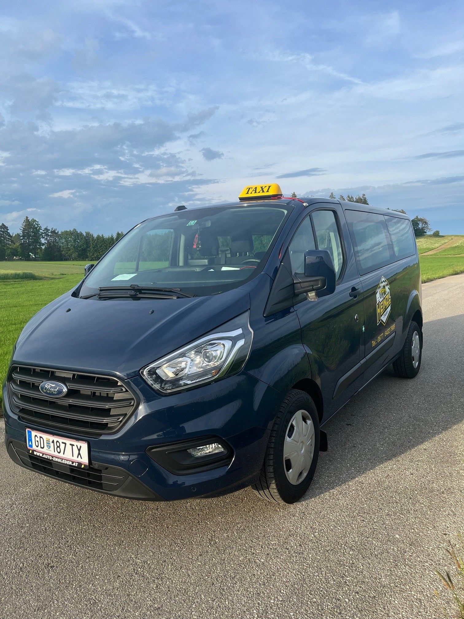Modrý taxík Ford stojí na vidieckej ceste pred zelenou lúkou a zamračenou oblohou.