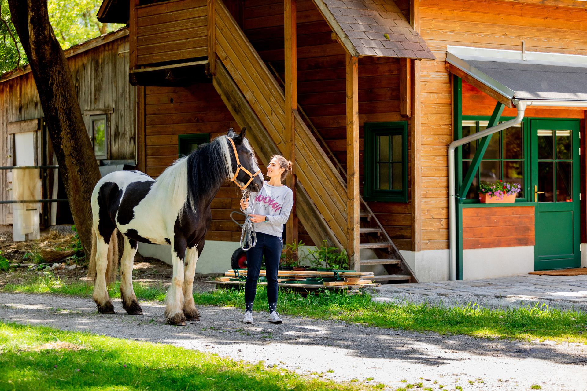 Žena stojí vedľa čiernobieleho koňa pred drevenou budovou.