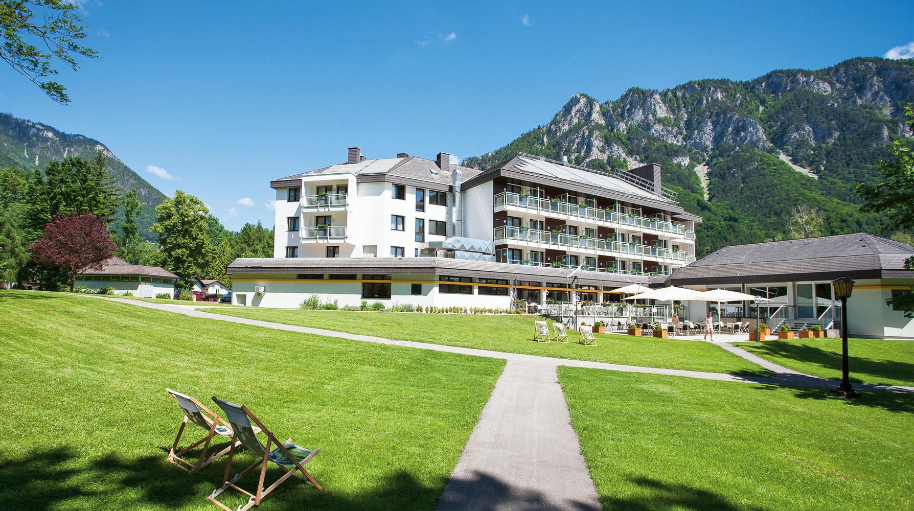 Parkhotel Hirschwang na pozadí hôr so zeleným trávnikom a ležadlami.