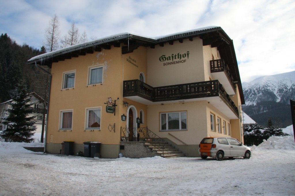 Žltá budova s nápisom "Gasthof Sonnenhof" na snehu, obklopená horami.