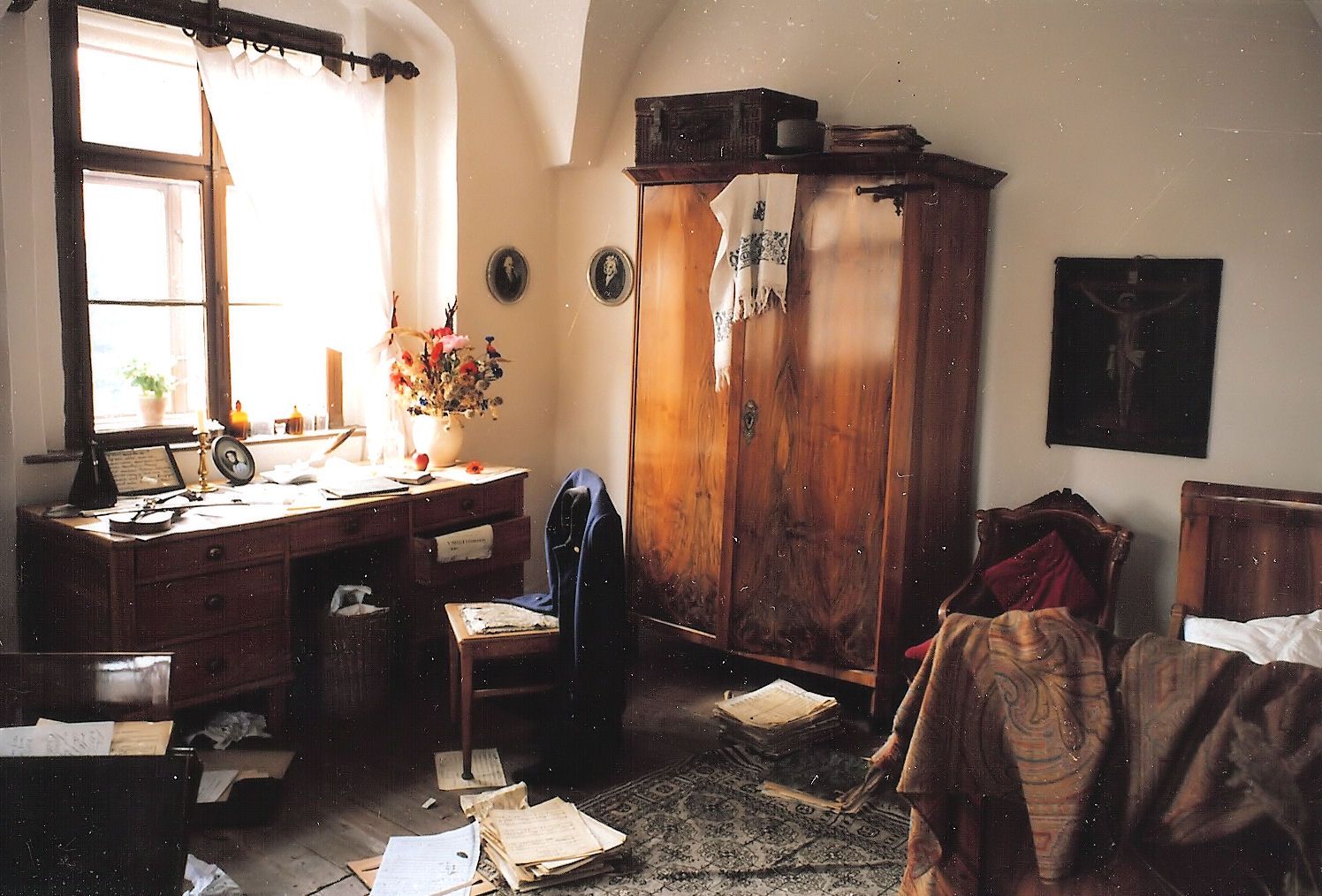 Historická izba s dreveným stolom, notami, skriňou a posteľou. Izba pôsobí neusporiadane a staromódne.