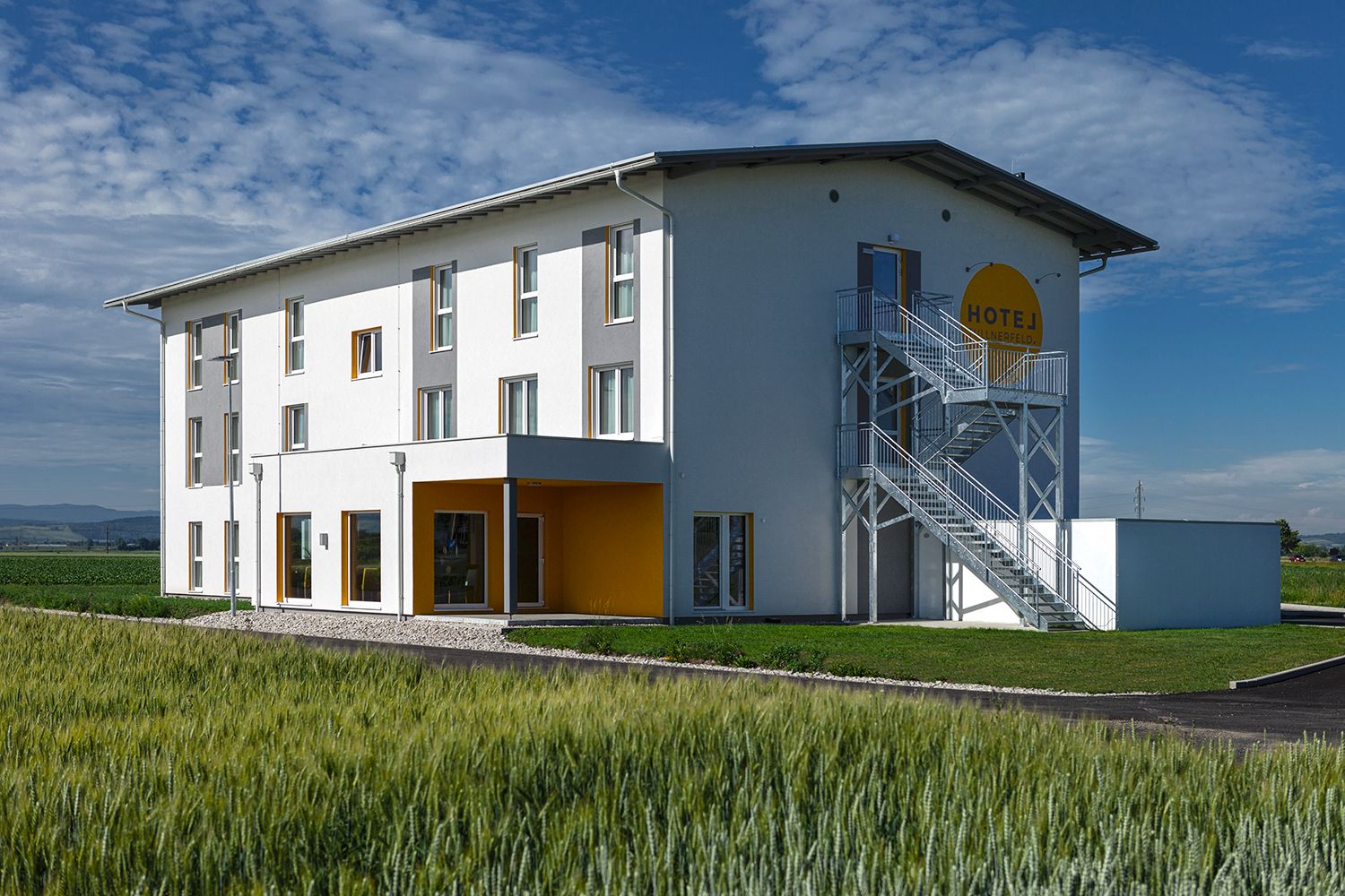 Exteriérový pohľad na moderný hotel s bielou fasádou a žltými akcentmi, obklopený poliami.