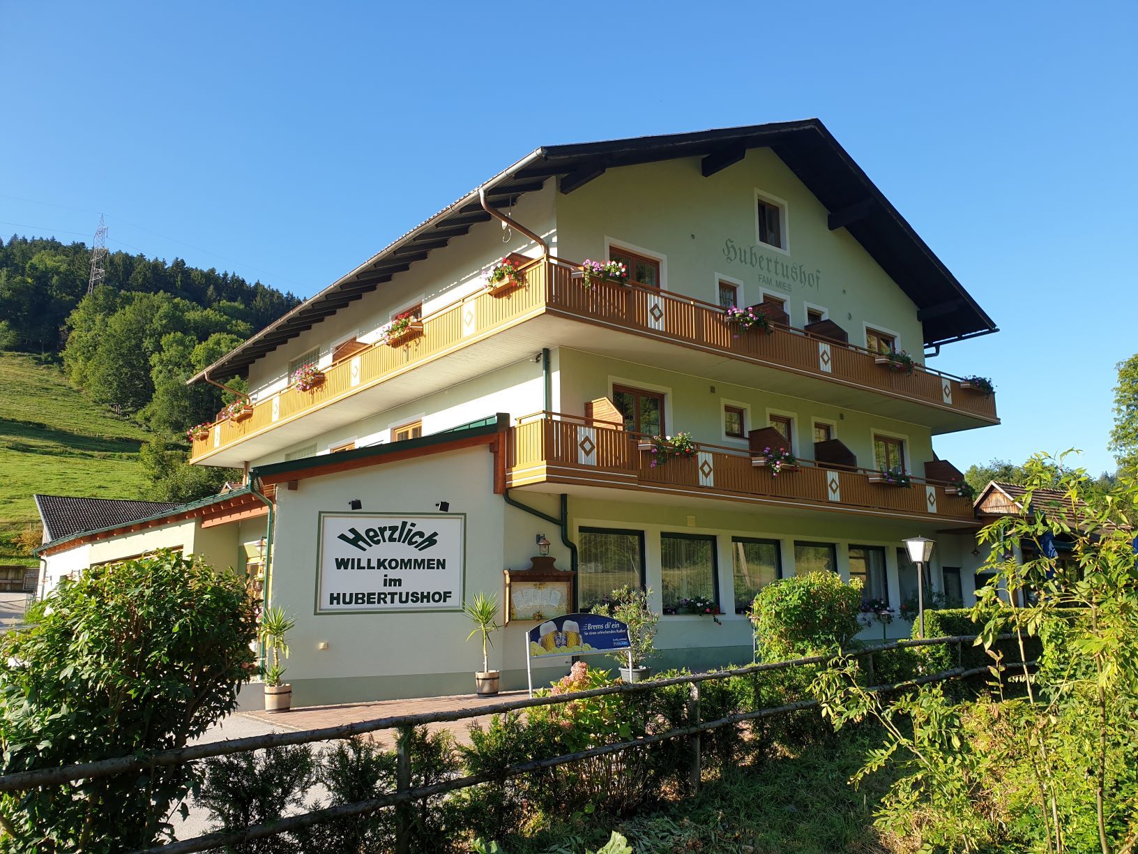 Tradičný hostinec Hubertushof s kvetmi na balkónoch, obklopený zelenou krajinou a modrou oblohou.