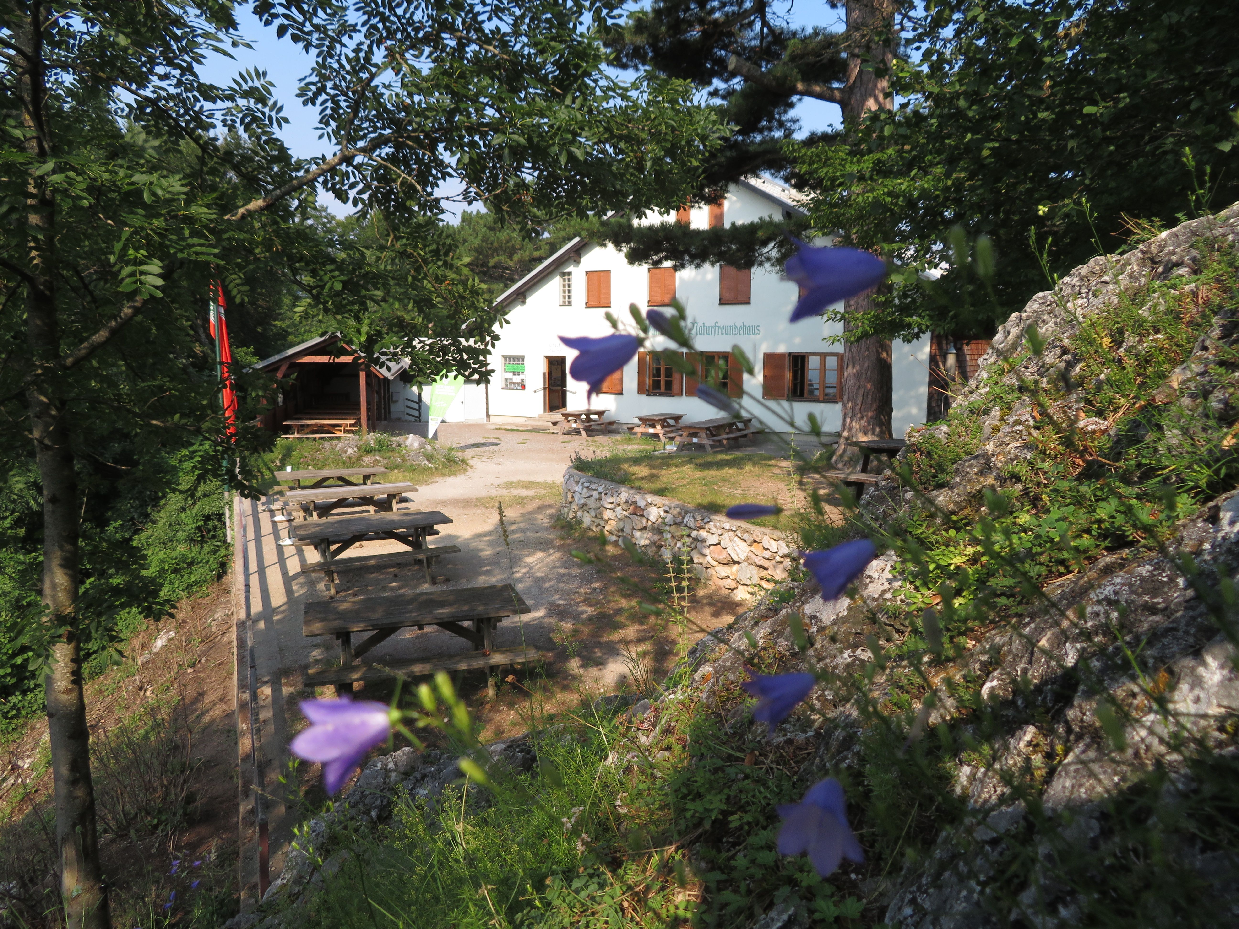 Biela budova s nápisom "Naturfreundehaus" obklopená stromami a kvetmi. V popredí vidno drevené lavičky a stoly.
