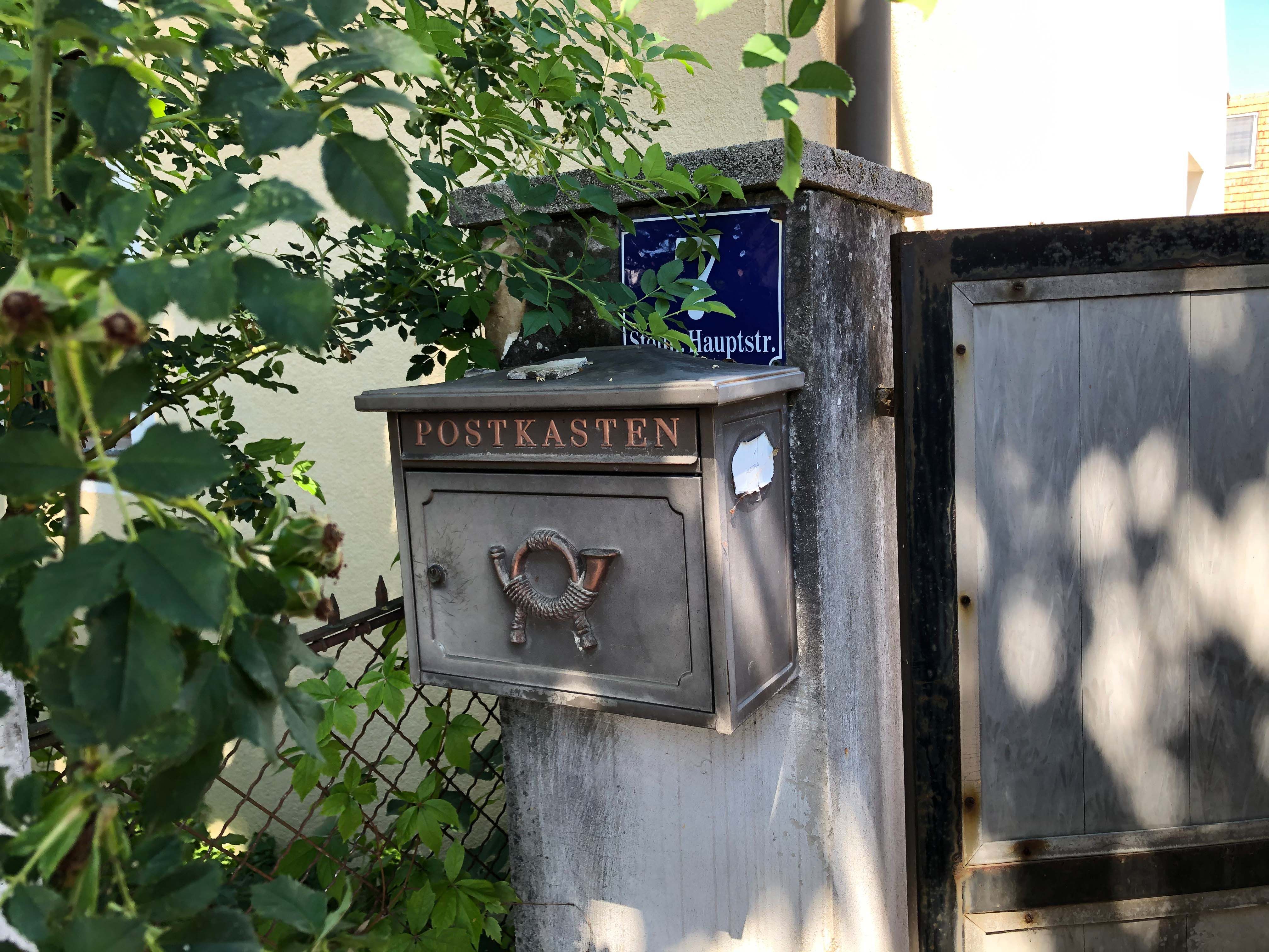 Stará poštová schránka s nápisom "Postbox" na múre vedľa brány, obklopená rastlinami.
